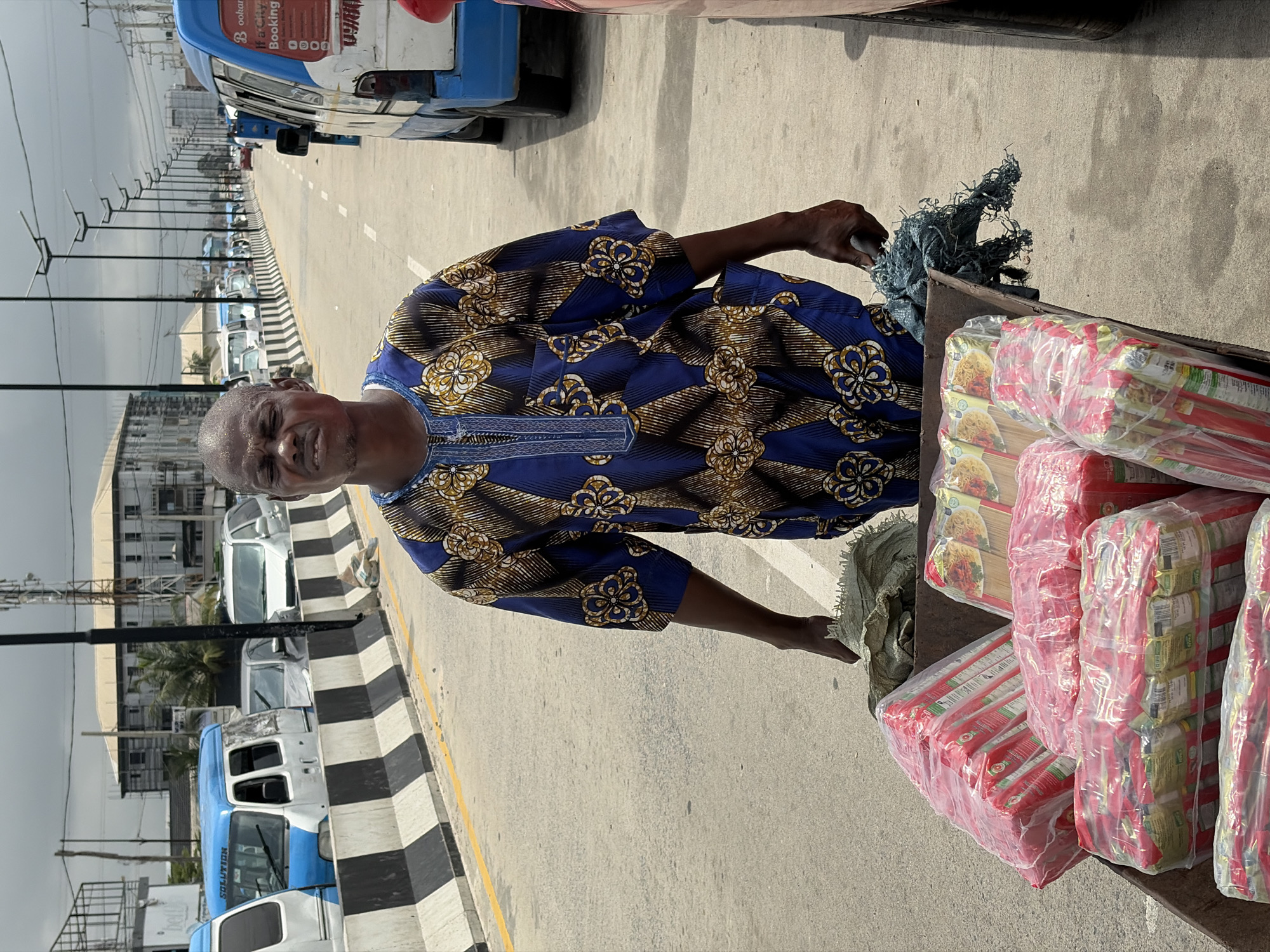 A portrait-oriented, vertical image shows a man standing next to a cart filled with packaged food items. The man is wearing a blue and gold patterned dashiki and appears to be looking slightly to his left, with a weathered expression. His right hand is extended and holding a piece of frayed rope. The cart is loaded with many rectangular packages, predominantly red and yellow, wrapped in clear plastic. These packages appear to be food items, possibly noodles or snacks, with images of food visible on some of the packaging.
Behind the man, a road is visible, lined with a concrete barrier painted with black and white stripes. Several vehicles, including what appears to be a white car and a blue bus with the word "SOLUTION" written on it, are parked along the road. In the background, there are buildings and utility poles, suggesting an urban or semi-urban environment. The lighting suggests it is daytime, and the sky appears overcast.
The scene is set in Igboefon, Nigeria, indicated by the location context. The overall impression is of a vendor in a busy area, possibly a market or a roadside stall, with his goods ready for sale. The man's posture and the presence of the cart suggest he is either preparing to sell, in the process of selling, or transporting his merchandise.