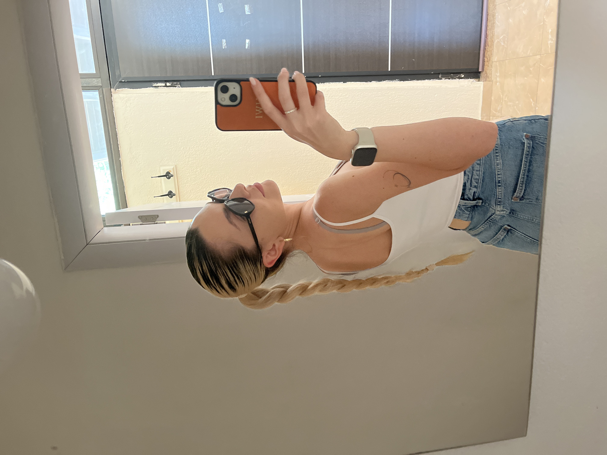 A woman is reflected in a mirror, seemingly taking a selfie. She is wearing a white tank top and light blue denim jeans. Her hair is blonde with darker roots and is styled in a long braid. She is wearing sunglasses. In her right hand, she holds a phone with an orange case, and on her wrist, she wears a light-colored watch. The reflection shows a room with a window, a door with brown panels, and beige tiled walls. The location appears to be indoors, possibly a bathroom.