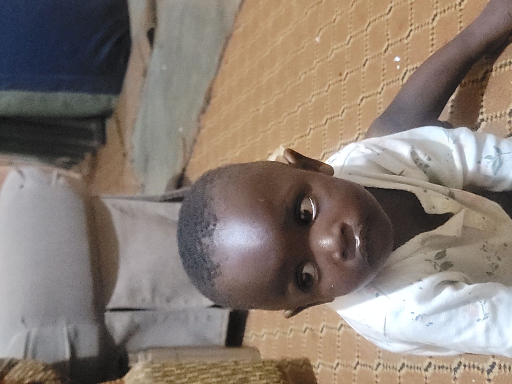 A young child, likely a toddler, is captured in a close-up portrait, seated on a brown, textured floor with a diamond pattern. The child has dark skin and short, dark hair. Their eyes are wide and looking off to the left, with a curious expression. There is a small amount of white residue around their mouth. The child is wearing a white long-sleeved shirt adorned with a subtle, light-colored floral pattern. To the left of the child, a portion of a woven basket is visible, and behind them, a blurred background shows what appears to be furniture or fabric in muted tones. The lighting suggests an indoor setting, possibly a home, in Barakin Akawo, Nigeria.