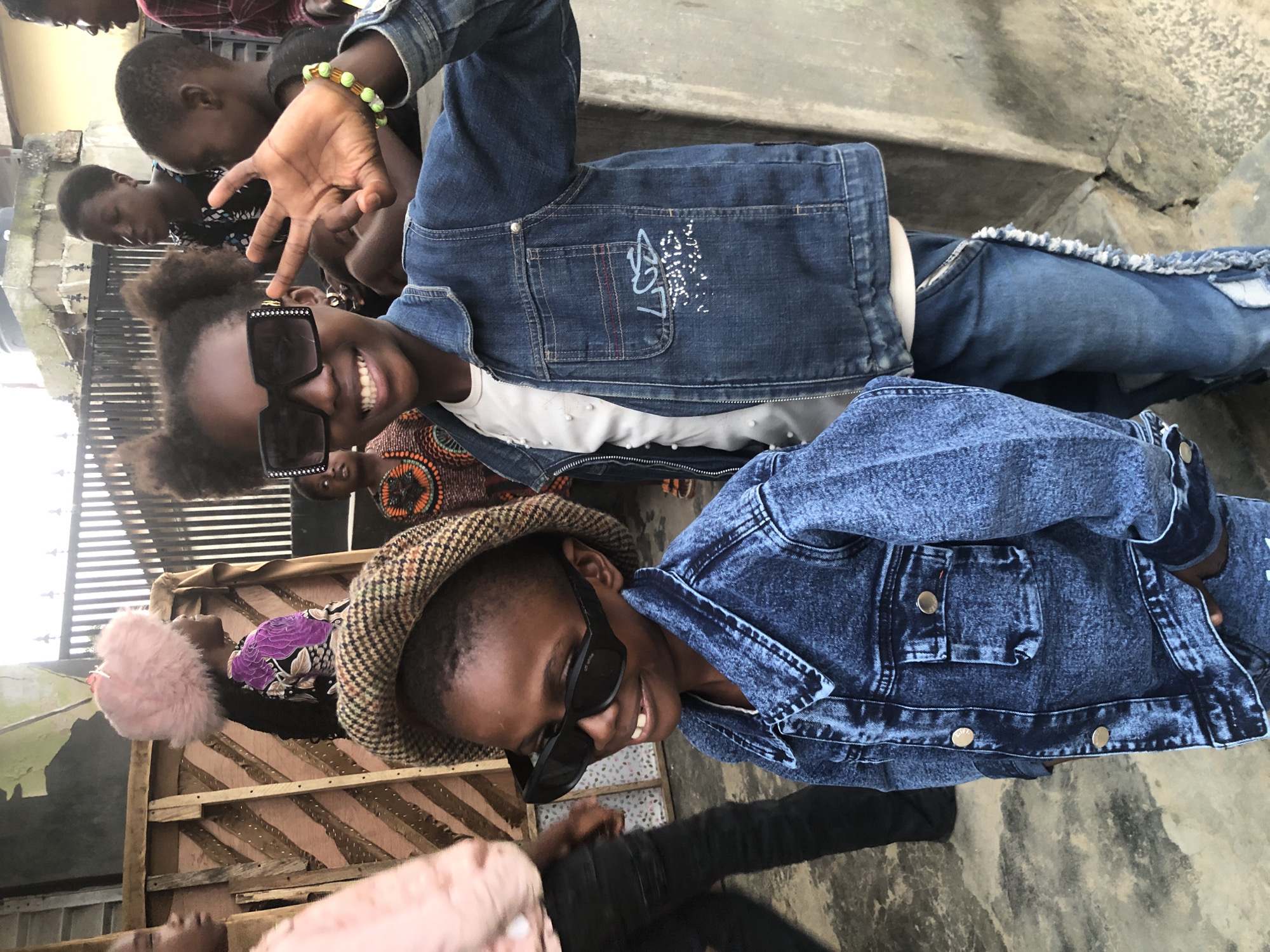 This is a medium shot of two young children, a girl and a boy, posing for a photo outdoors in Ado, Nigeria. The girl, in the foreground and on the right, wears large, square sunglasses with rhinestones, a blue denim jacket over a white t-shirt, and jeans. She has her right hand raised, showing three fingers, and is smiling broadly. A beaded bracelet adorns her left wrist. To her left and slightly behind her, the boy wears a brown and beige plaid cap, large black sunglasses, and a blue denim jacket. He is also smiling and looking towards the camera. Several other children are visible in the background, some partially obscured. The setting appears to be a street or courtyard, with a rough concrete wall on the right and a wooden structure with slatted panels on the left. A metal fence is visible behind the children. The ground is made of concrete with visible patches and cracks. The overall lighting suggests it is daytime.