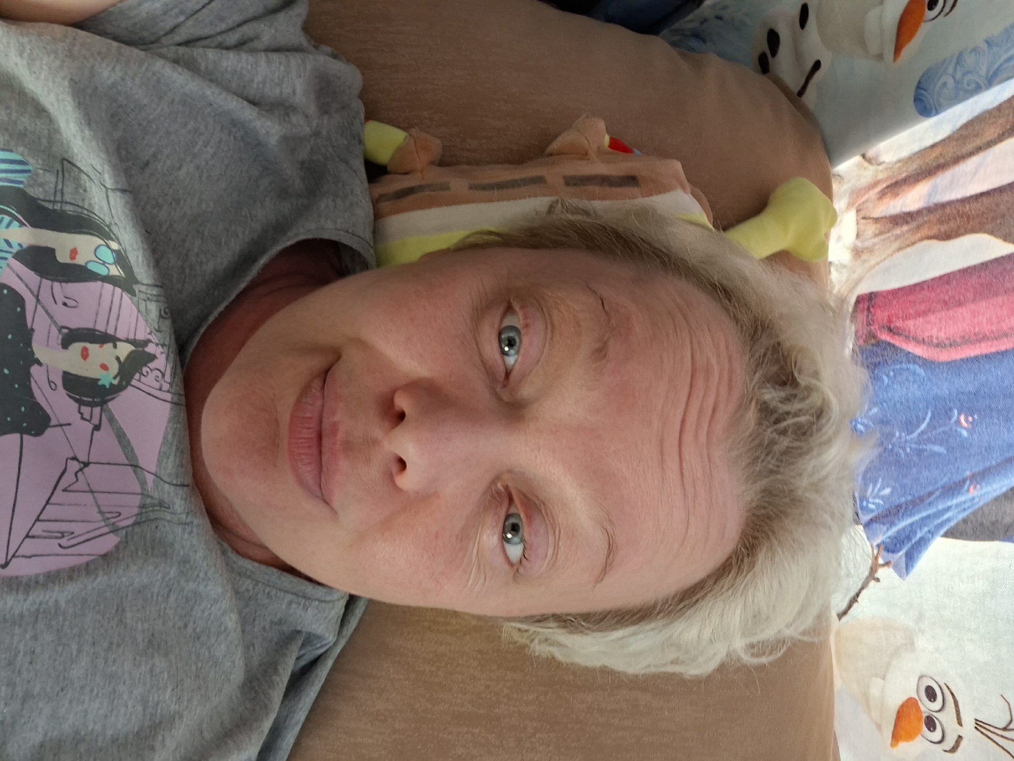 A close-up, overhead shot shows a person with fair skin and short, light blonde hair. The person is lying down, looking directly at the camera. Their eyes are open and appear to be light blue. They have visible wrinkles on their forehead and around their eyes. The person is wearing a grey t-shirt with a graphic print of two cartoon-like women. A stuffed toy, possibly a Spongebob Squarepants character, is visible next to their head. In the background, a blanket or sheet with animated characters, including what appears to be Olaf from Frozen, is visible. The setting appears to be indoors, possibly a bed or sofa. No specific location in Novoilinsky, Russia can be determined from the image.