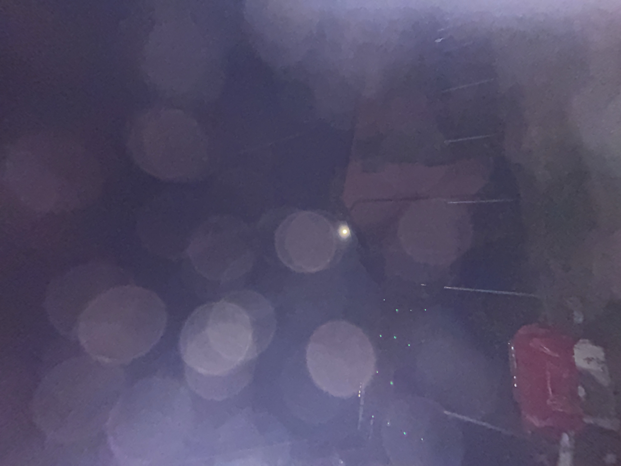 The image appears to show a blurry scene, possibly taken through a window covered in raindrops. There are multiple out-of-focus circles, likely representing droplets reflecting light. A red object is visible towards the right side of the image. There is no information in the image to determine the location in the city of Sofia, Bulgaria. No people, subjects, actions, or interactions can be identified due to the lack of clarity. The environment is vaguely suggested to be outdoors at night or in dim light.