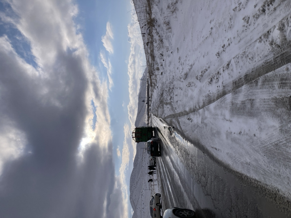 The image depicts a winter scene on a mountainous road in Churmian, Pakistan. The two-lane asphalt road runs through a snow-covered landscape under a partly cloudy sky with visible sunlight breaking through. The road surface is wet and slushy in places, with significant snow accumulation and tire tracks on the shoulders and unpaved areas to the right.
Vehicles are present on the road: a partial view of a white vehicle is in the bottom left foreground. Further down the left lane, a silver sedan is stopped on the shoulder. Adjacent to this silver sedan, a group of approximately five to six individuals is gathered, standing and interacting. In the right lane, a dark-colored sedan follows a large, green heavy vehicle, possibly a dump truck, with a tall, open cargo bed appearing to contain hay or vegetation. Both vehicles are traveling away from the viewer.
The surrounding environment consists of snow-covered hills and mountains extending into the distance. Sparse, dry vegetation protrudes from the snow. In the far distance on the right, power lines are visible.