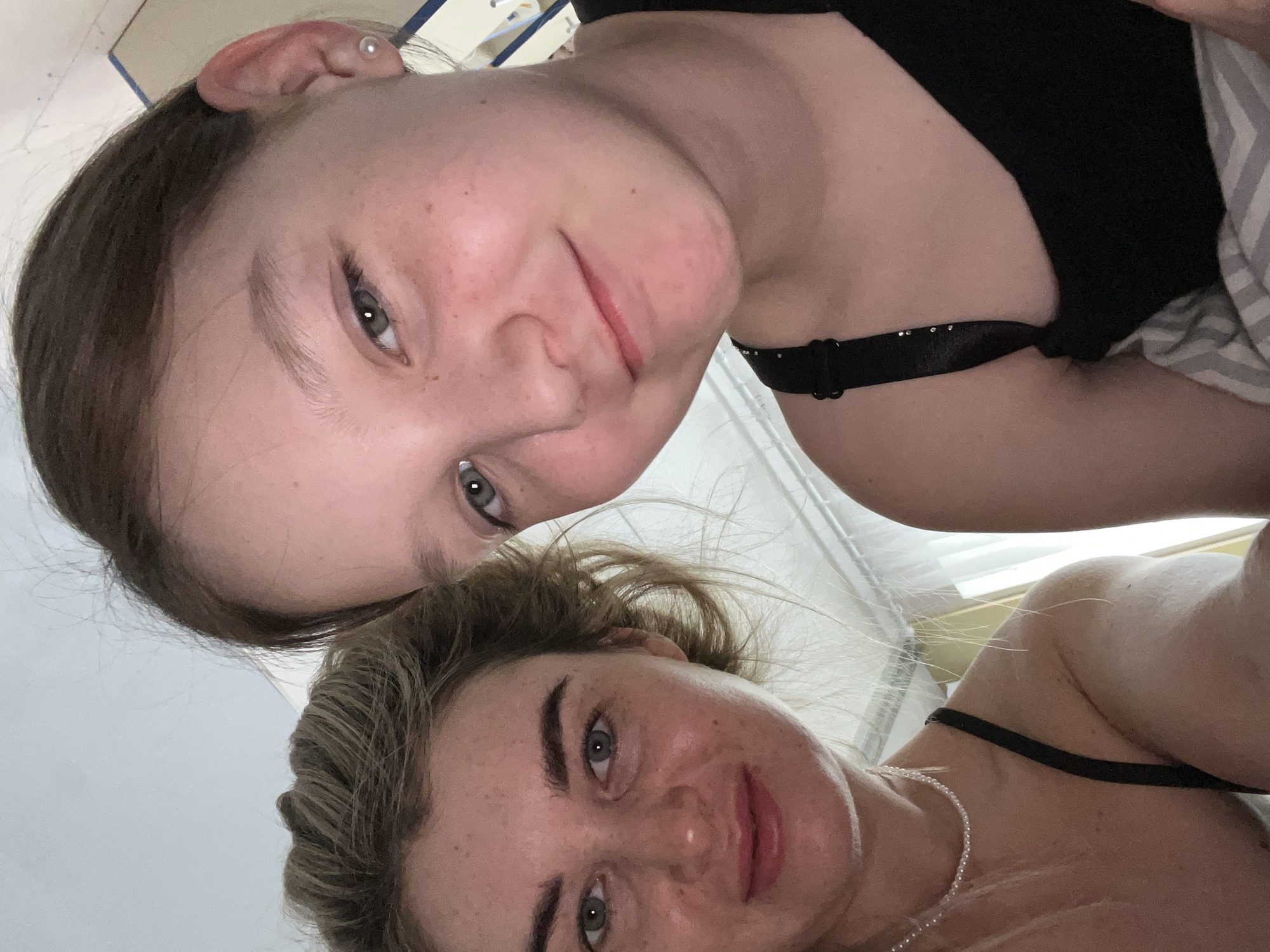 A close-up selfie of two women is presented. The upper woman has light brown hair, fair skin with freckles, and grey eyes. She is wearing a black top with a decorative strap. The lower woman has blonde hair, fair skin with freckles, and light-colored eyes. She wears a black top and a delicate pearl necklace. They are indoors, with a window behind them. The location is unknown and cannot be inferred to be Chaikovsky, Russia.