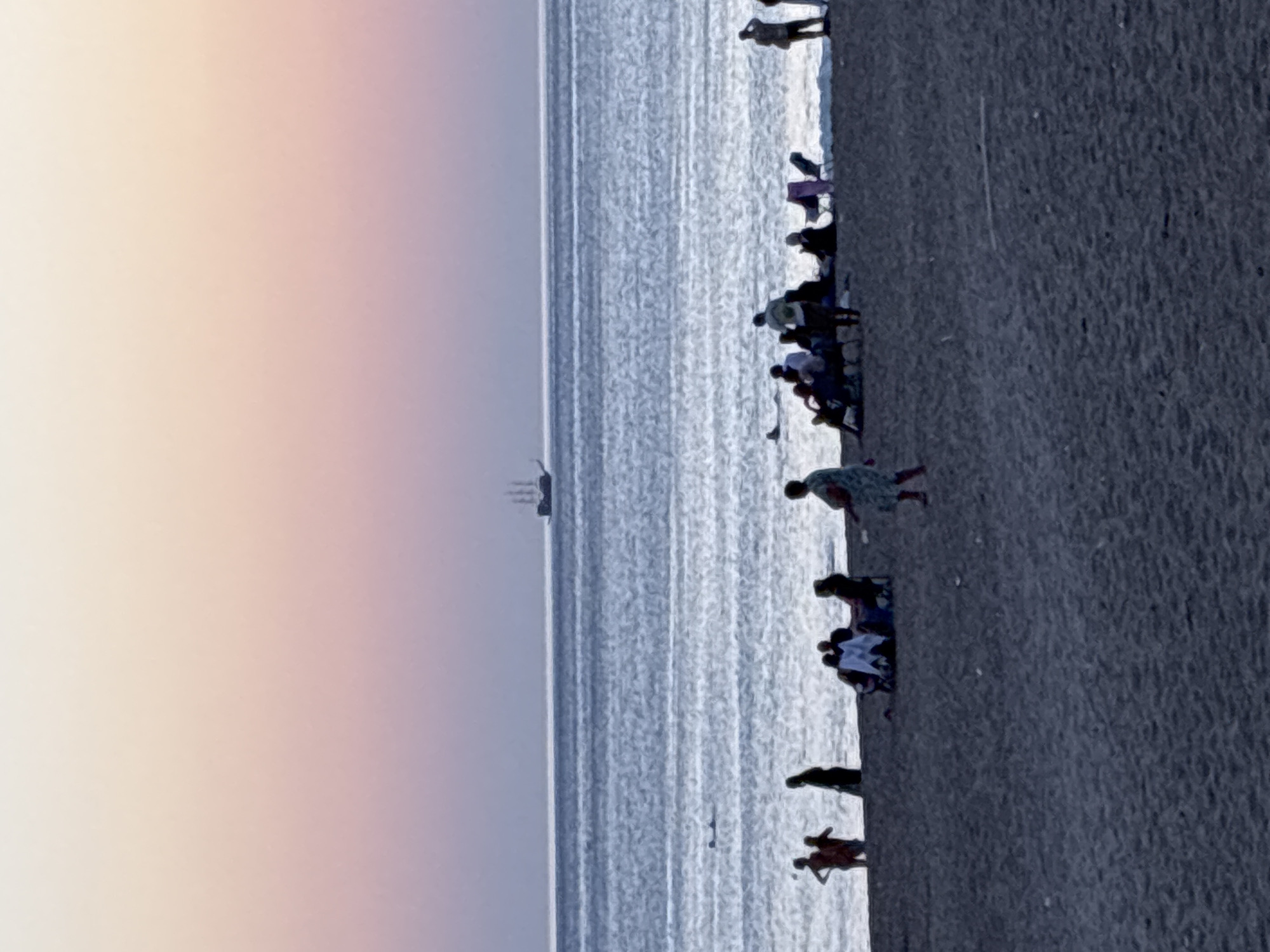 The image depicts a beach scene at dusk in Alanya, Turkey. In the distance, a tall ship with multiple sails is visible on the calm sea. The sky displays a gradient of pastel colors, transitioning from pale yellow at the top to pink and purple near the horizon. The sea is a deep blue with shimmering reflections. On the sandy beach, several groups of people are gathered. In the foreground and midground, silhouetted figures can be seen sitting on chairs, lying on blankets, and standing. Some individuals are near the water's edge, while others are further up the beach. The overall atmosphere is serene and relaxed, characteristic of an evening on the Mediterranean coast.