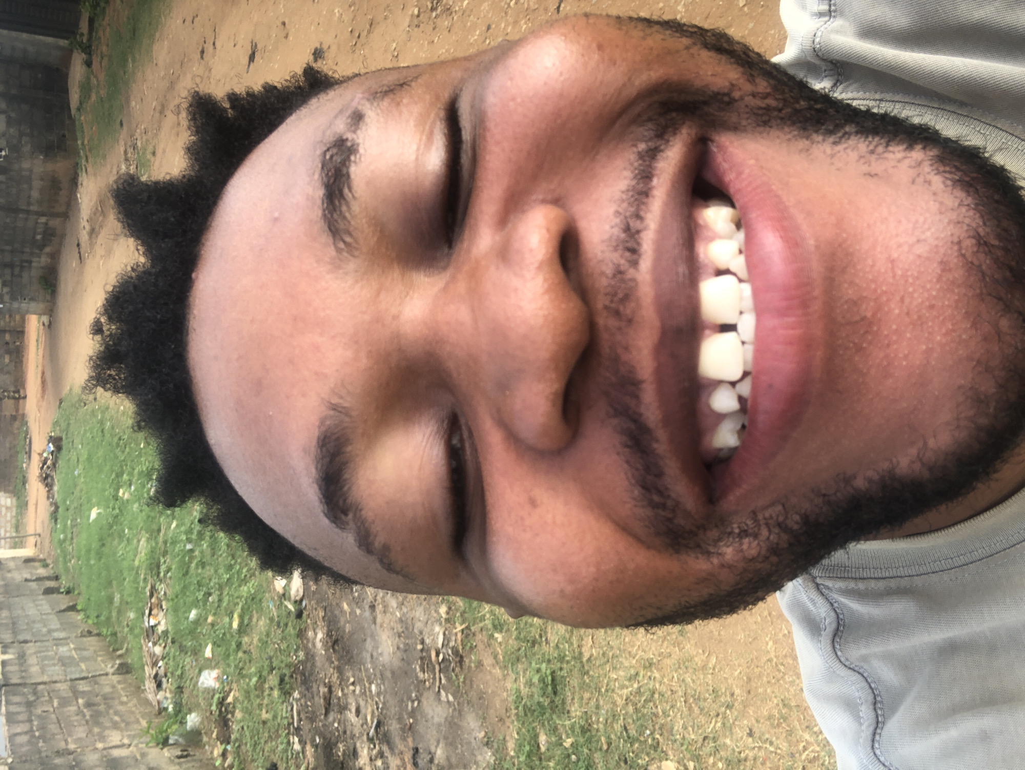 A close-up, rotated image shows a man with dark skin, closed eyes, and a broad smile revealing his teeth. He has short, dark curly hair and a beard. He is wearing a light gray t-shirt. The background consists of a dirt or gravel path with patches of green grass and trash. A low brick wall is visible in the distance. The location is likely outdoors in an urban or semi-urban environment. The exact city and country are unknown.