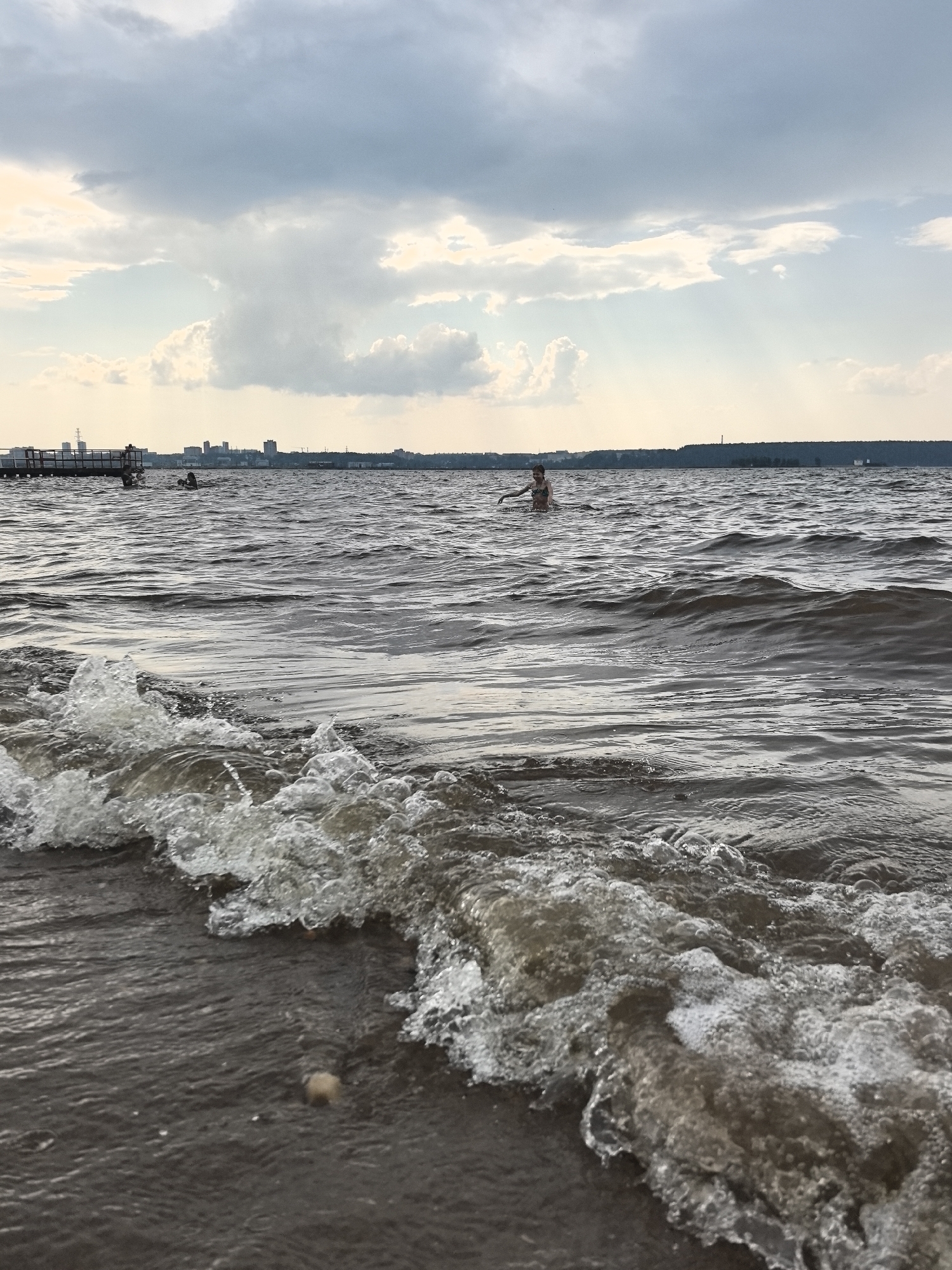 The image depicts a natural body of water, likely a river or lake, with a shoreline in the foreground. The water is choppy, with small waves breaking on the sandy or pebbly shore. In the distance, a cityscape is visible on the horizon, suggesting an urban environment. A pier or dock extends into the water on the left side of the image, with a few figures on it. Further out in the water, a person is swimming. The sky is cloudy, with patches of sunlight breaking through. The overall impression is one of a recreational setting, possibly a public beach or park. The specific location in the city of Perm, Russia, is suggested by the presence of the cityscape on the horizon, which may include identifiable Perm landmarks.