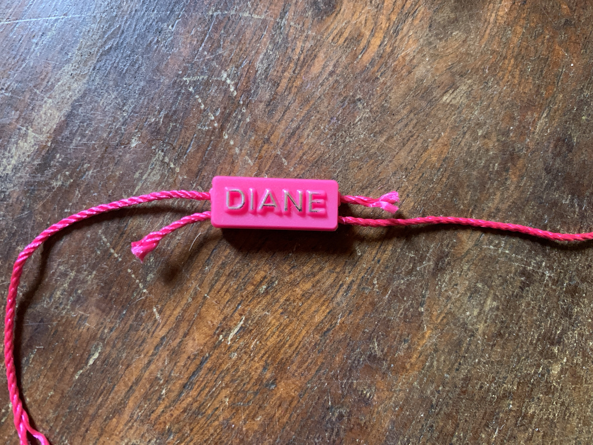 The image displays a close-up view of a personalized pink charm on a braided pink string, resting on a textured wooden surface.
**Subjects:** The primary subject is a small, bright pink, rectangular charm. A thin, braided pink string passes through the charm. The background is a worn, wooden surface.
**Activity or event taking place:** No activity or event is taking place. The objects are stationary, likely laid out for display or observation.
**Notable details:**
The pink charm is rectangular with rounded edges. On its surface, the name "DIANE" is embossed in silver-colored capital letters, which show some wear or fading, revealing the underlying pink plastic in places. The charm has holes on either side through which the pink string is threaded. The string itself is a vibrant fuchsia pink, appearing braided or twisted, and seems to be made of a synthetic material. Simple knots secure the string on both sides of the charm, and the ends of the string beyond the knots are frayed.
The background is a rustic wooden surface, possibly a table or floor, characterized by a rich brown hue with visible grain patterns, scratches, and lighter areas suggesting wear and age. The lighting is soft and even, without harsh shadows.
**Visible text:** The name "DIANE" is clearly visible in raised, silver-colored letters on the pink charm.
**Location cues:** The image does not contain any visual cues to confirm the location context of Santa Ana, Venezuela.