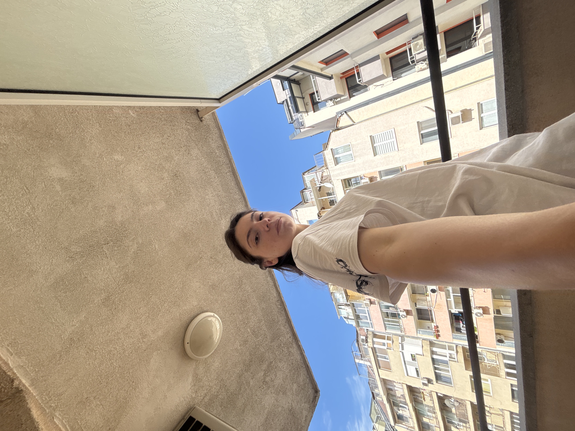 This is a first-person perspective, overhead shot of a young woman lying down, likely on a balcony or rooftop, in Varna, Bulgaria. The woman is wearing a white t-shirt with a graphic print. She is looking up towards the camera with a neutral expression.
The scene is an urban setting, with apartment buildings visible in the background. The buildings are multi-story structures with balconies and windows, painted in a light yellow and beige color palette with orange accents. The sky is a clear, bright blue, suggesting a sunny day, likely mid-morning or early afternoon.
In the foreground, part of a textured wall and a light fixture are visible, along with a portion of the woman's arm. A black metal railing runs across the frame, separating the woman from the view of the buildings. The overall impression is of a relaxed, casual moment captured outdoors on a clear day in a city. There is no visible text in the image.