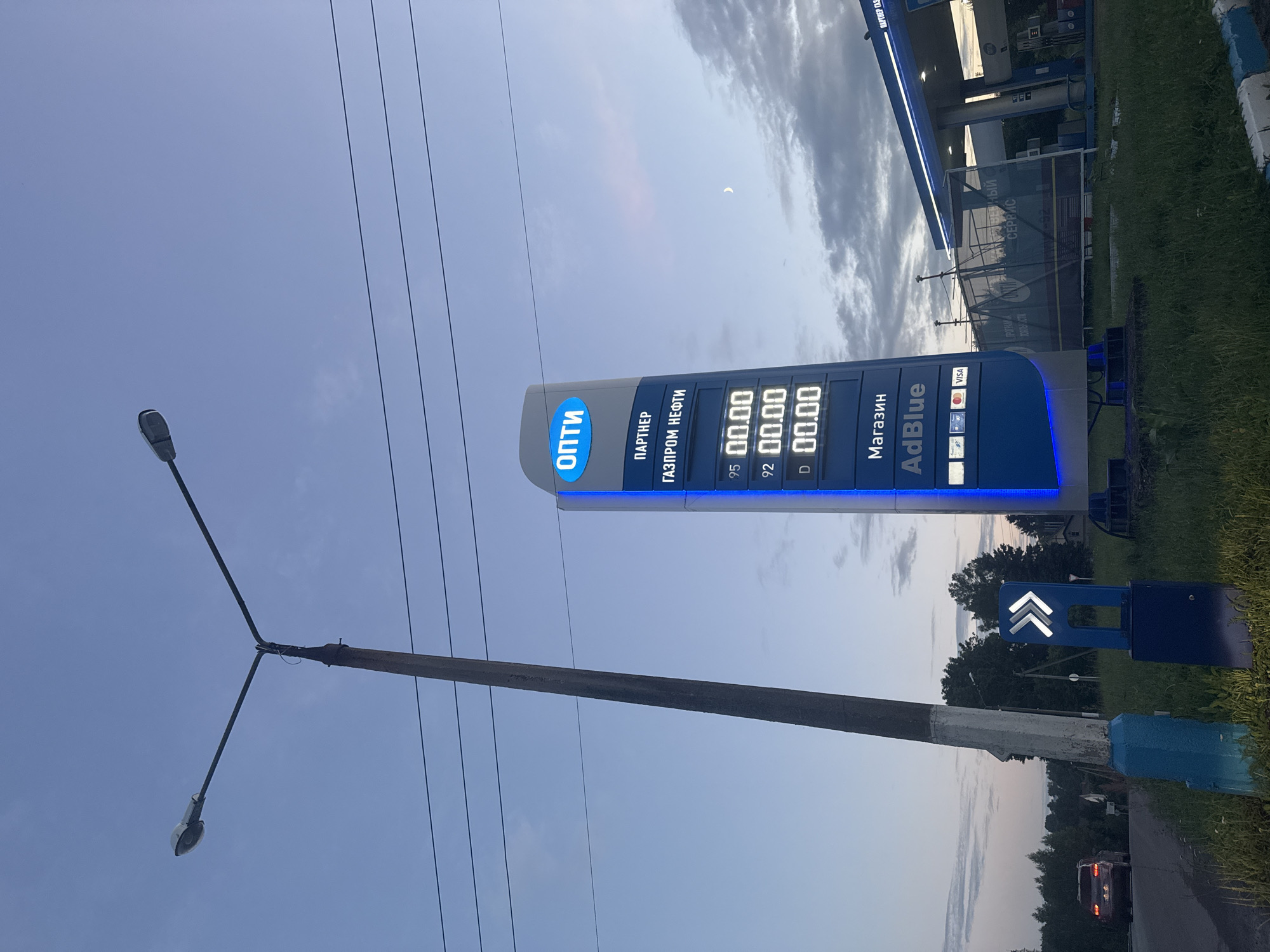 This image depicts a view of a gas station in the city of Vereya, Russia, during dusk. A prominent blue and white gas price display tower is visible on the right, showing prices for different fuel types, with "ОПТИ" and "ГАЗПРОМ НЕФТИ" branding. Below the fuel prices, "AdBlue" is advertised. To the left of the price display, a tall concrete utility pole with a street lamp stands, casting a shadow on the surrounding grass. A dark-colored car is seen driving away on a road in the background on the left. The sky is a soft blue with scattered clouds, and a crescent moon is visible. Further to the right, a portion of the gas station's canopy is visible, illuminated by blue lights. The overall environment appears to be a semi-urban or rural roadside setting.