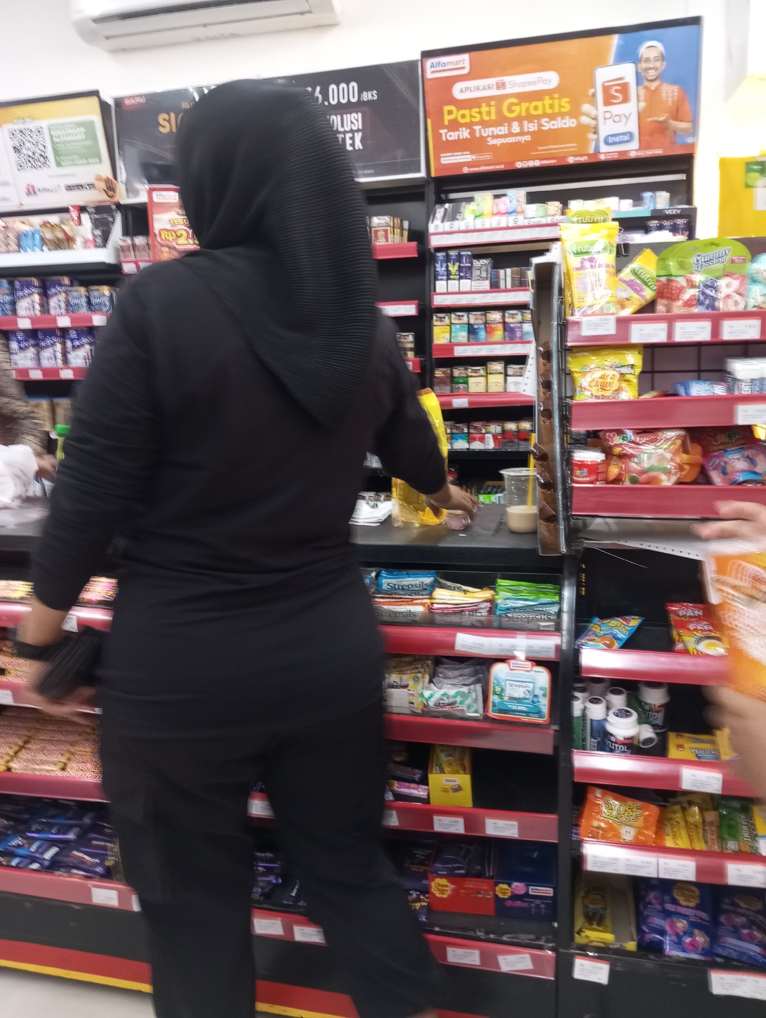 An interior shot captures a person from behind, wearing a black hijab and black clothing, as they stand in an aisle of an Alfamart convenience store. The store features red shelving units stocked with various products, including packaged snacks, drinks, toiletries, and cigarette cartons. A prominent advertising banner for ShopeePay is displayed at the top of the aisle. The floor appears to be tiled. The lighting is artificial and typical of a retail environment. The individual's focus is directed towards the shelves, suggesting they are browsing or shopping.
