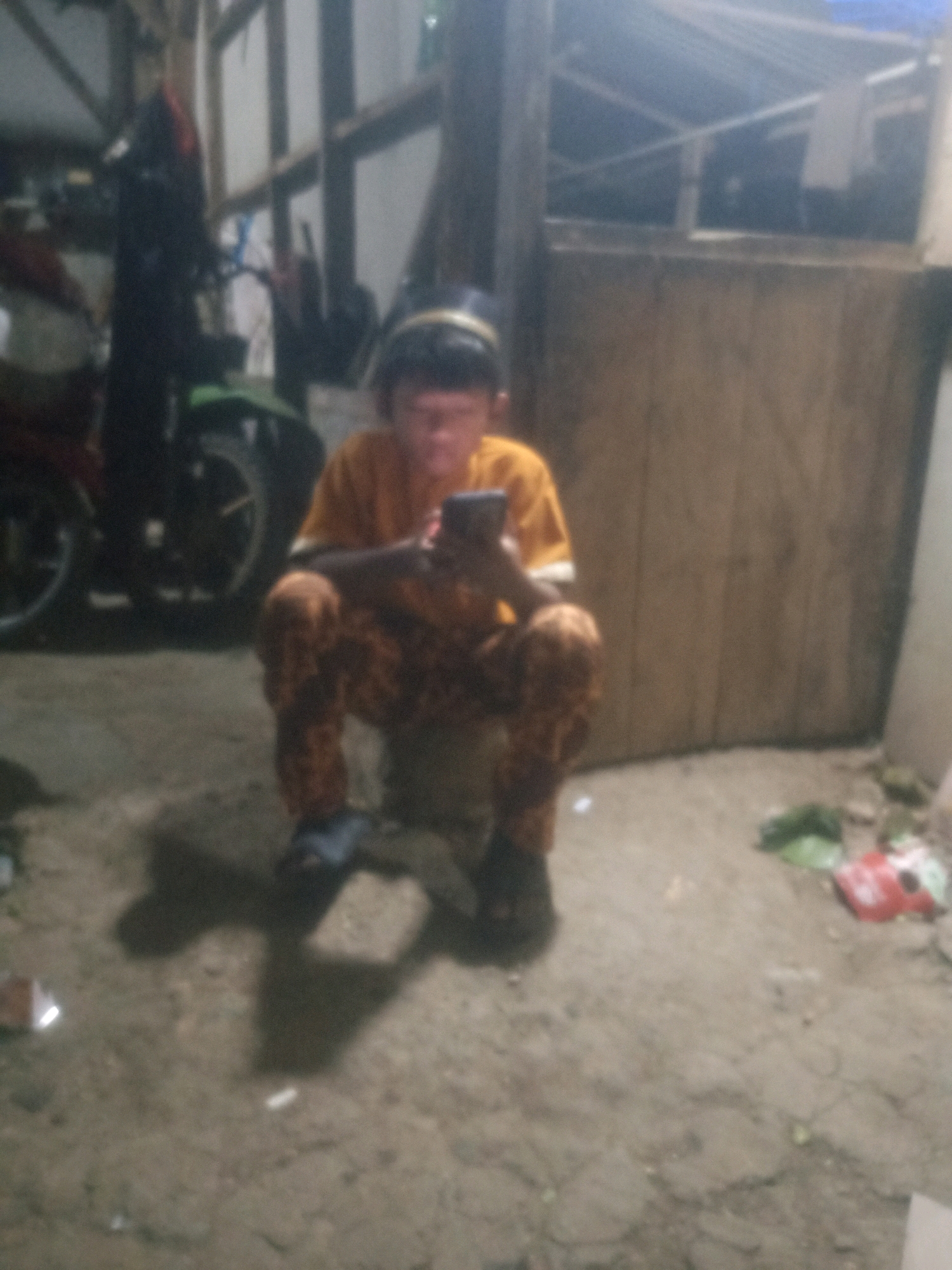 A person is crouched down in an outdoor, possibly rural, environment, holding a smartphone and looking at its screen. They are wearing a bright yellow t-shirt and patterned pants. In the background, a motorcycle and wooden structures are visible. The ground is dirt and appears to have some debris. The lighting suggests it may be evening or indoors with artificial light. The image is low quality and blurry, making precise identification of features difficult.
