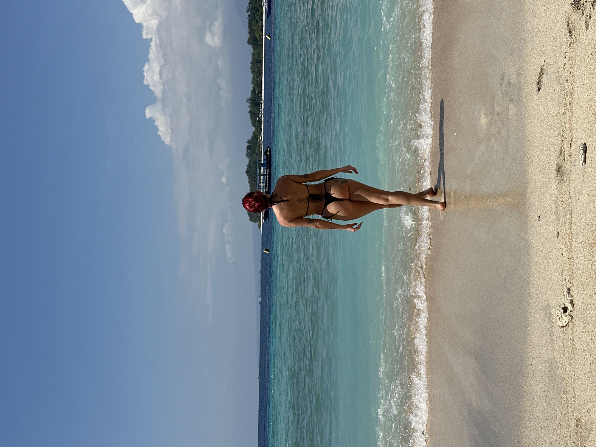 The image shows a person standing on a beach in Unknown, Indonesia. The person, viewed from the back, has fair skin and red hair tied up in a bun. She is wearing a black bikini and appears to be walking toward the water. The water is turquoise and clear, meeting the sandy beach with gentle waves. The sky is blue with fluffy white clouds. In the distance, trees are visible on the coastline. There are footprints in the sand.