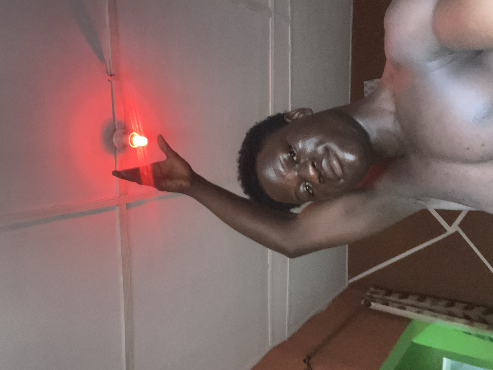 A shirtless, dark-skinned man is partially visible in the frame. He is facing the camera with his head tilted, and his skin appears glossy. His left arm is extended, with his hand positioned near a light fixture on the ceiling. The light fixture emits a red glow. The background consists of a white wall with vertical molding. The photo appears to have been taken in an interior setting. There is no information available from the image to determine if this photo was taken in Mosafejaw, Nigeria.