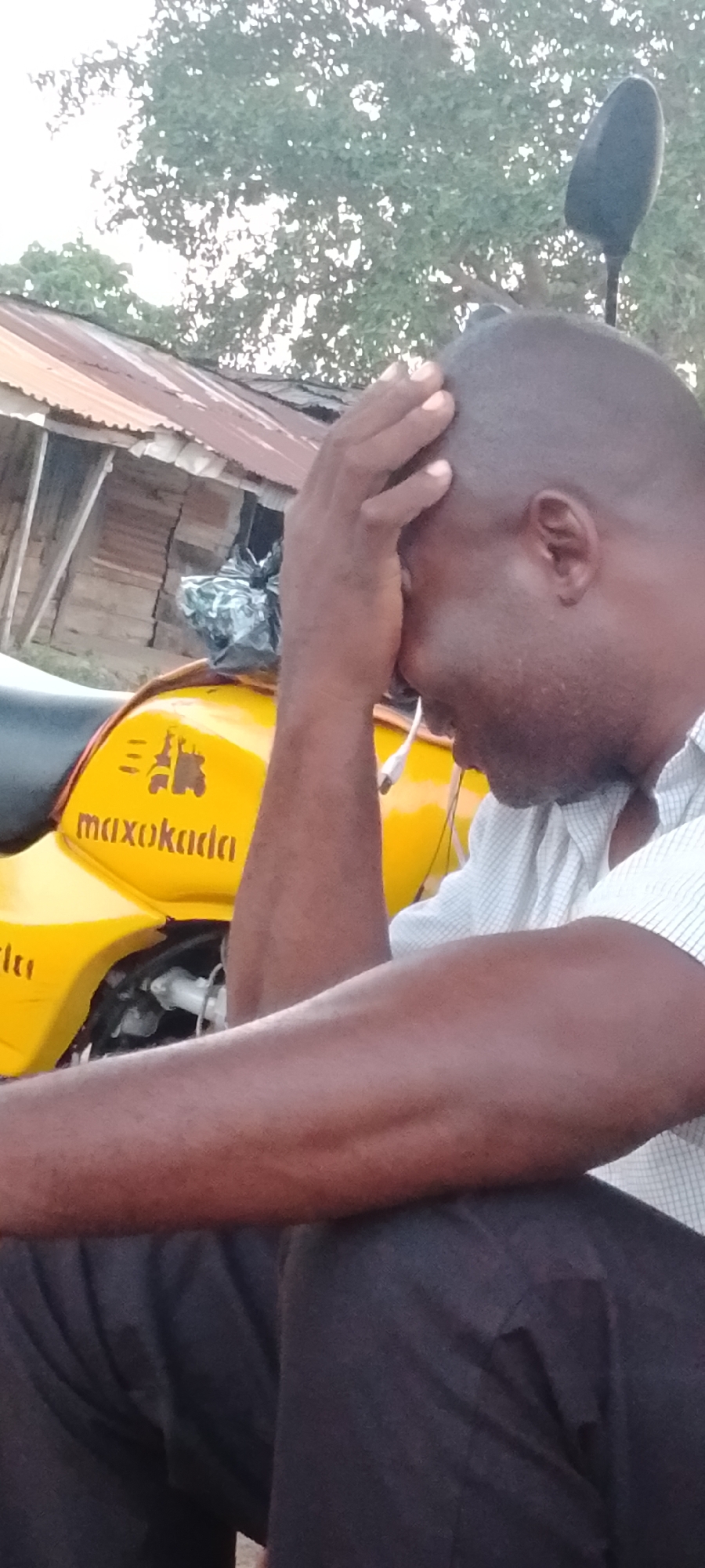 A man is seated next to a yellow motorcycle in Adankolo, Nigeria. He is dark-skinned and has his hand on his head. The motorcycle has the word "maxokada" on the side. A building with a corrugated metal roof is in the background. A tree is also visible.