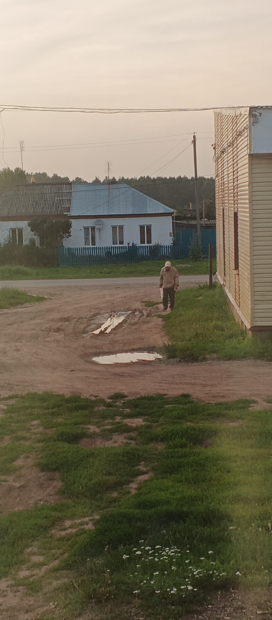 A man wearing a light-colored hat and jacket walks on a dirt road in front of a residential building and a long, light-yellow building. A blue fence is visible in front of the residential building. The sky is overcast and the scene appears to be in a rural or suburban setting. There are puddles of water on the dirt road. Trees and foliage are visible in the background. The building on the right is a long, corrugated metal structure. The residential building has a gray and blue roof. Power lines are visible in the sky.