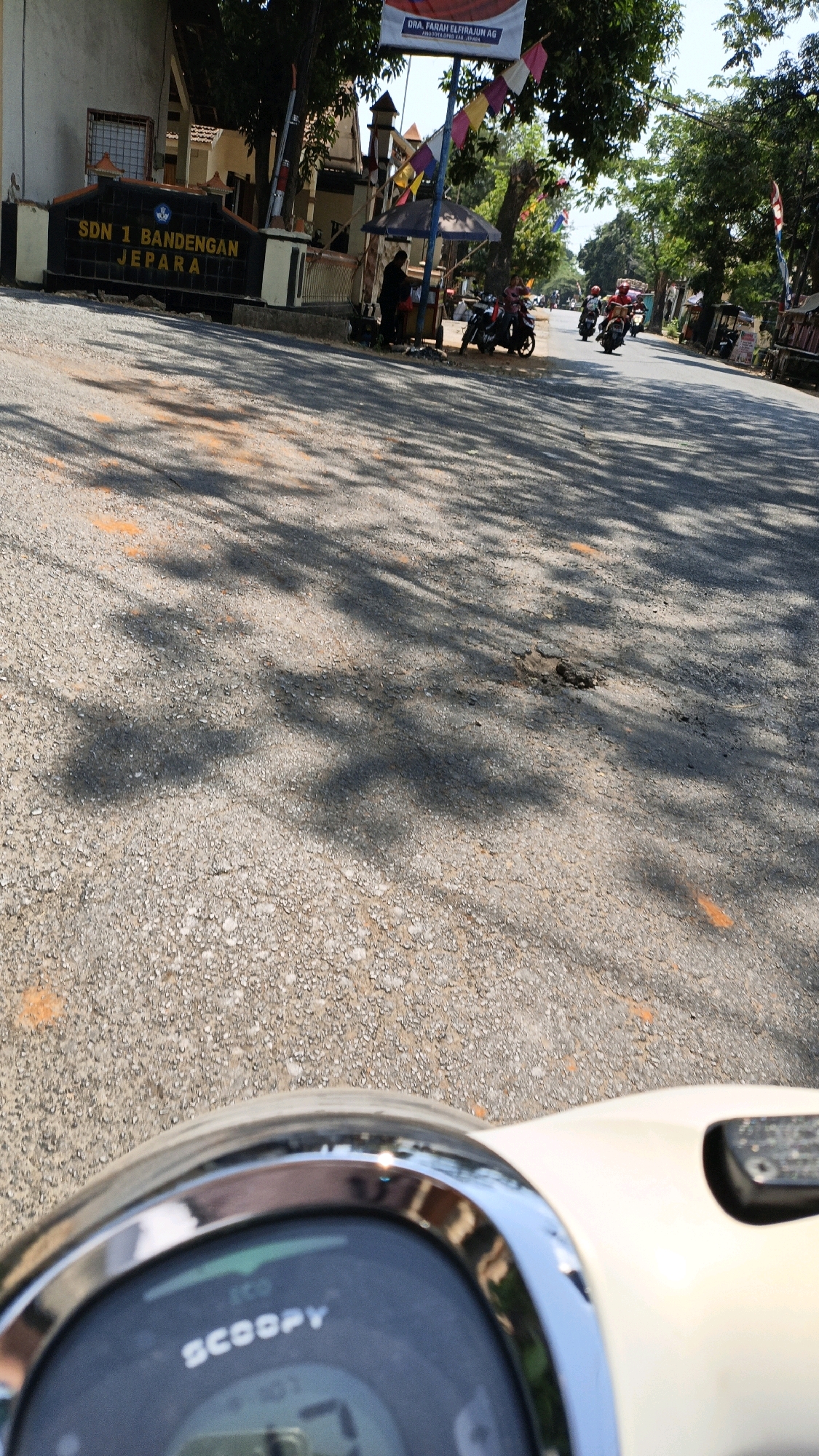 The image depicts a street scene in Bandengan, Indonesia, in front of SDN 1 Bandengan Jepara. The foreground shows the dashboard of a scooter, with the "SCOOPY" logo visible. The road surface is asphalt with visible gravel and some orange markings. Tree shadows cast dappled patterns across the road. In the mid-ground, a sign for "SDN 1 BANDENGAN JEPARA" is prominent. Several motorcycles are parked or in motion on the road. A few individuals are present near the school entrance, some standing and others on motorcycles. Colorful flags are strung across the area. The background shows trees and buildings, suggesting a sunny day. The overall environment appears to be a school entrance area on a public road.
