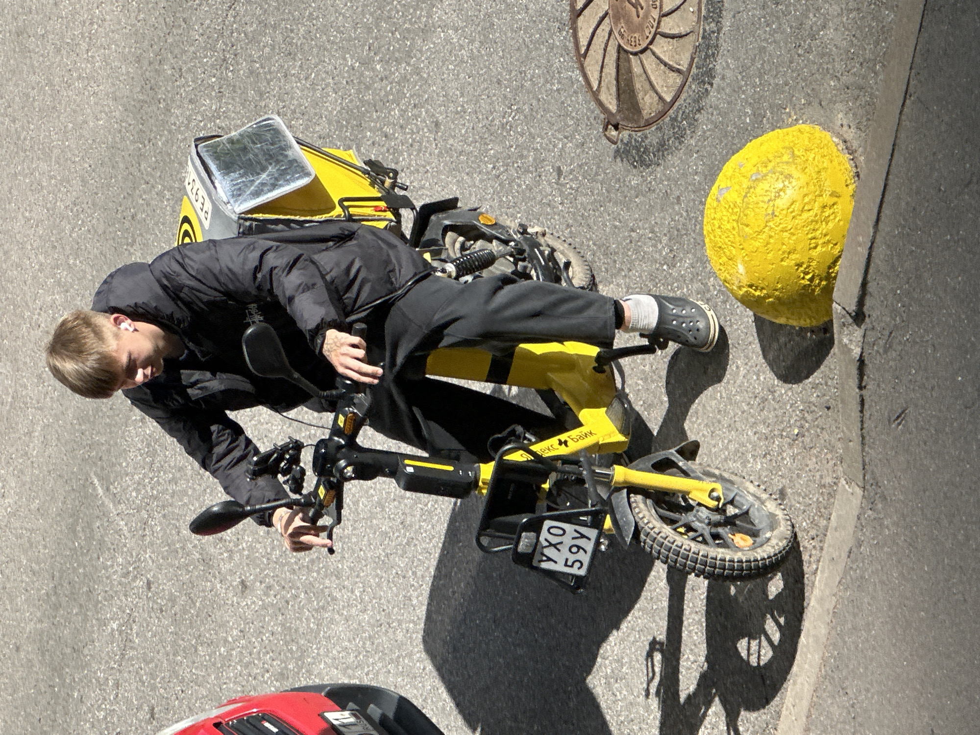 A young man with blonde hair and white earbuds is riding a yellow electric scooter on a paved city street in Murino, Russia. He is wearing a black puffer jacket, black sweatpants, white socks, and dark crocs. The scooter has a large yellow storage box on the back and a license plate that reads "УХО 59У". A red vehicle is partially visible on the left side of the image. A yellow concrete hemisphere is in the foreground on the right, and a manhole cover is also visible. The ground is asphalt, and there are shadows cast by the scooter and the man.