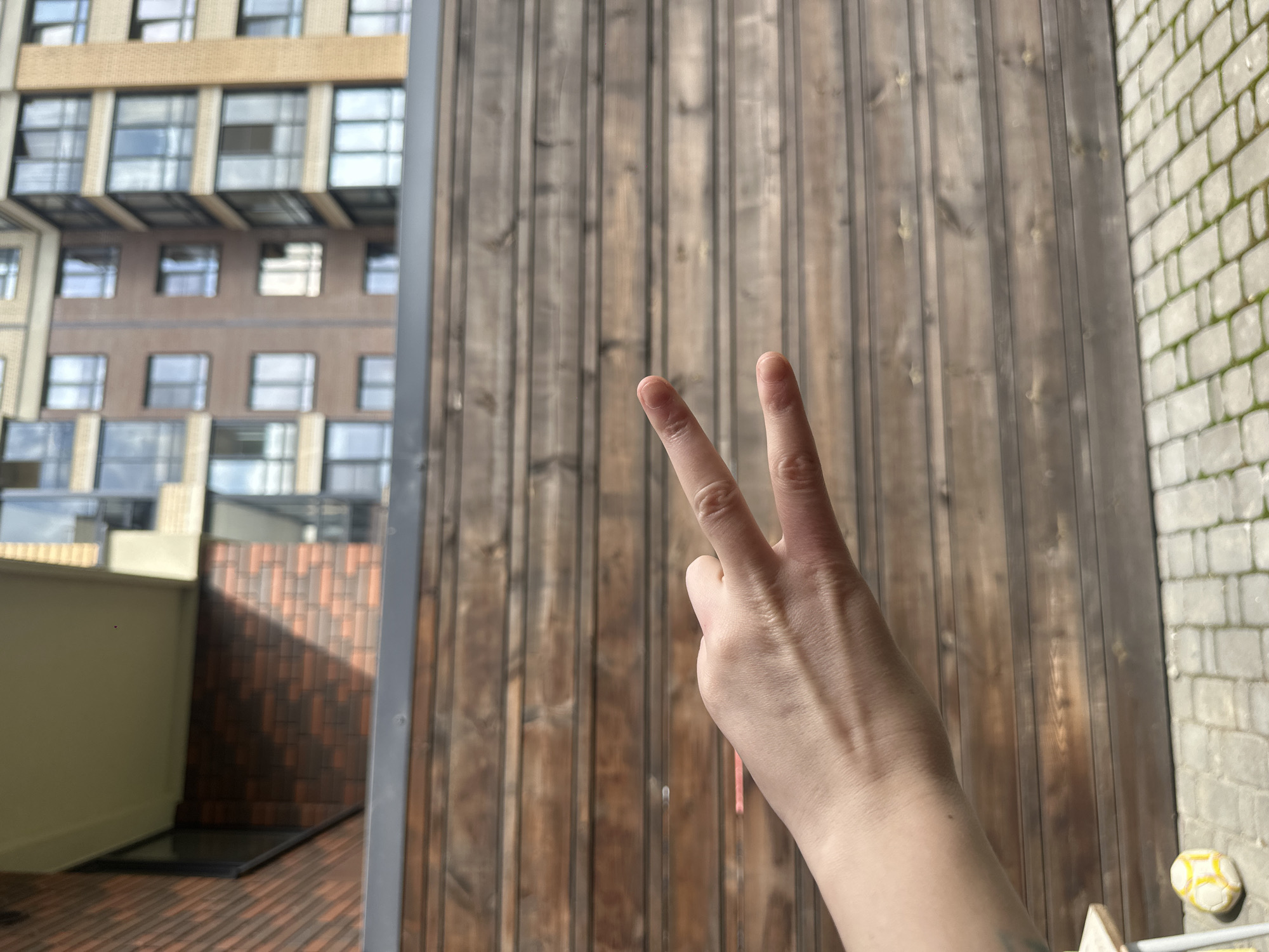 A hand is held up in the foreground, with the index and middle fingers extended in a peace sign gesture against a background of a wooden fence. Behind the fence, a brick building with multiple windows is visible. The ground in front of the fence is paved with light-colored stones. A small, yellow and white ball is visible on the paving stones in the lower left corner of the image.