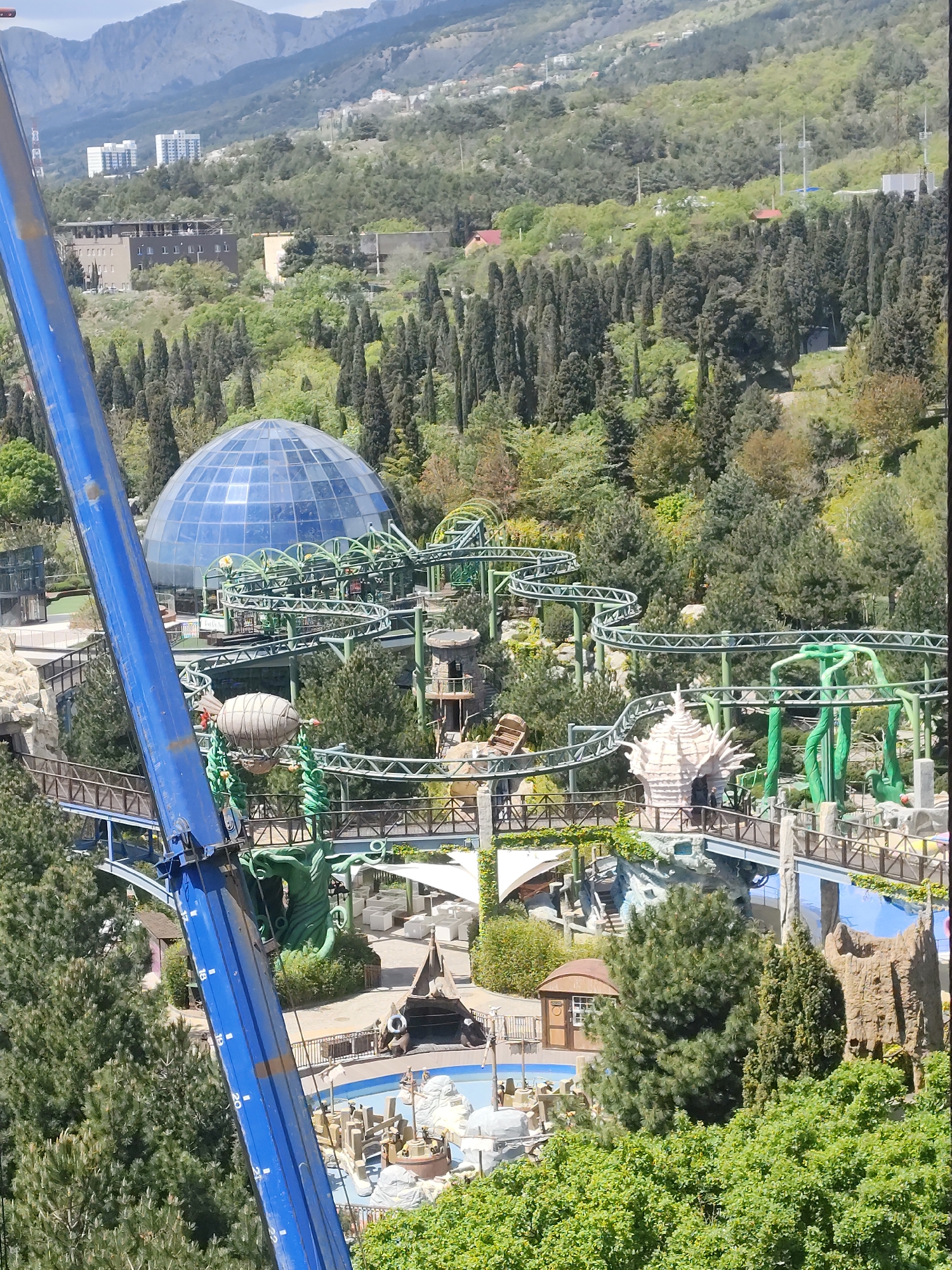 The image shows a section of a theme park located in Симеизский поселковый совет, Ukraine. A blue crane arm is prominently visible in the foreground, partially obscuring the view. The park features a complex of rides and attractions, including a green-painted roller coaster that winds through the trees, a large blue dome-shaped structure, a water feature with faux rock formations, and various fantasy-themed structures and decorations. The environment includes a mix of evergreen and deciduous trees, and several buildings can be seen in the distance on the slopes of a mountain range.