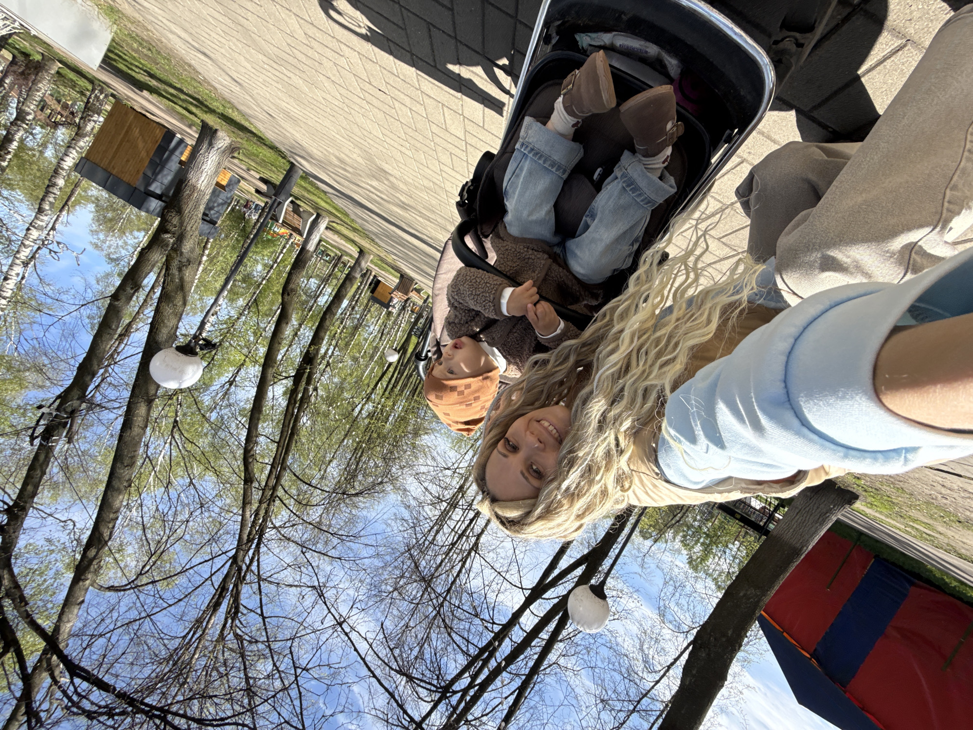 The image shows a woman holding a stroller with a baby inside. They are in a park or outdoor area with trees, and there is a building in the background. The photo was taken in the city of Шебунино, Russia.
