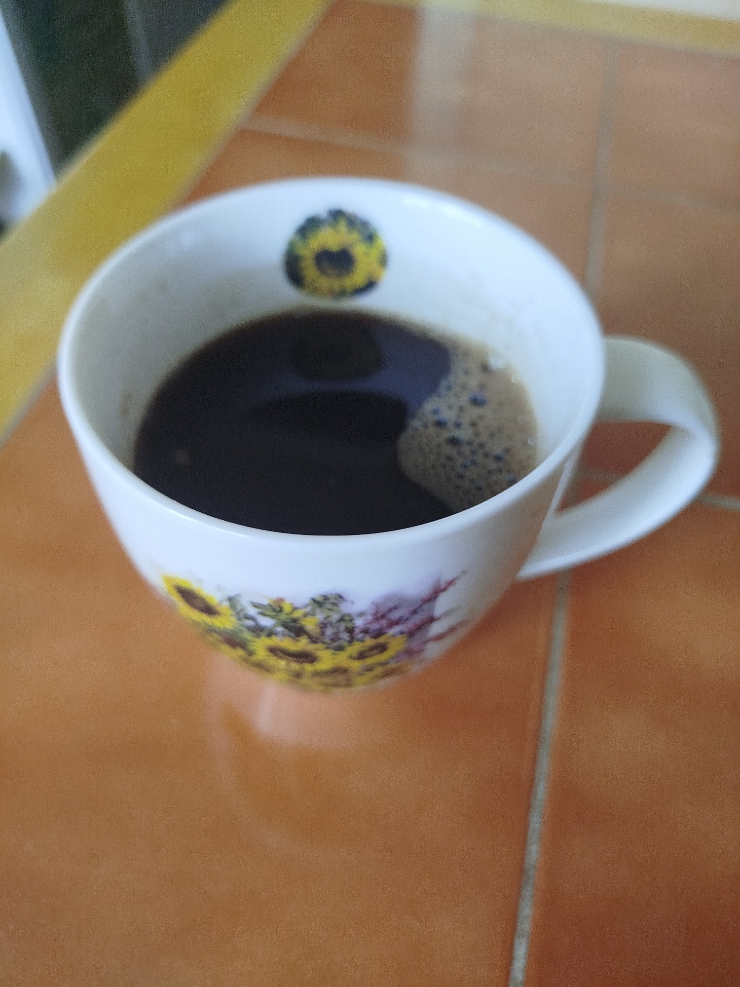 A close-up, overhead shot captures a white ceramic coffee mug filled with dark coffee, resting on a tiled surface. The mug features a decorative pattern of sunflowers and possibly other flowers. The coffee has a frothy surface with small bubbles. The tiled surface is a warm, earthy orange with grout lines visible. A yellow object, possibly a placemat or a strip of fabric, is partially visible in the upper left corner of the image. The lighting suggests an indoor environment, possibly a kitchen or dining area. The image does not contain any people or visible interactions. The scene is static and focused on the coffee mug and its immediate surroundings. The location of Tolbitsa, Russia, is not depicted or implied in the image itself.