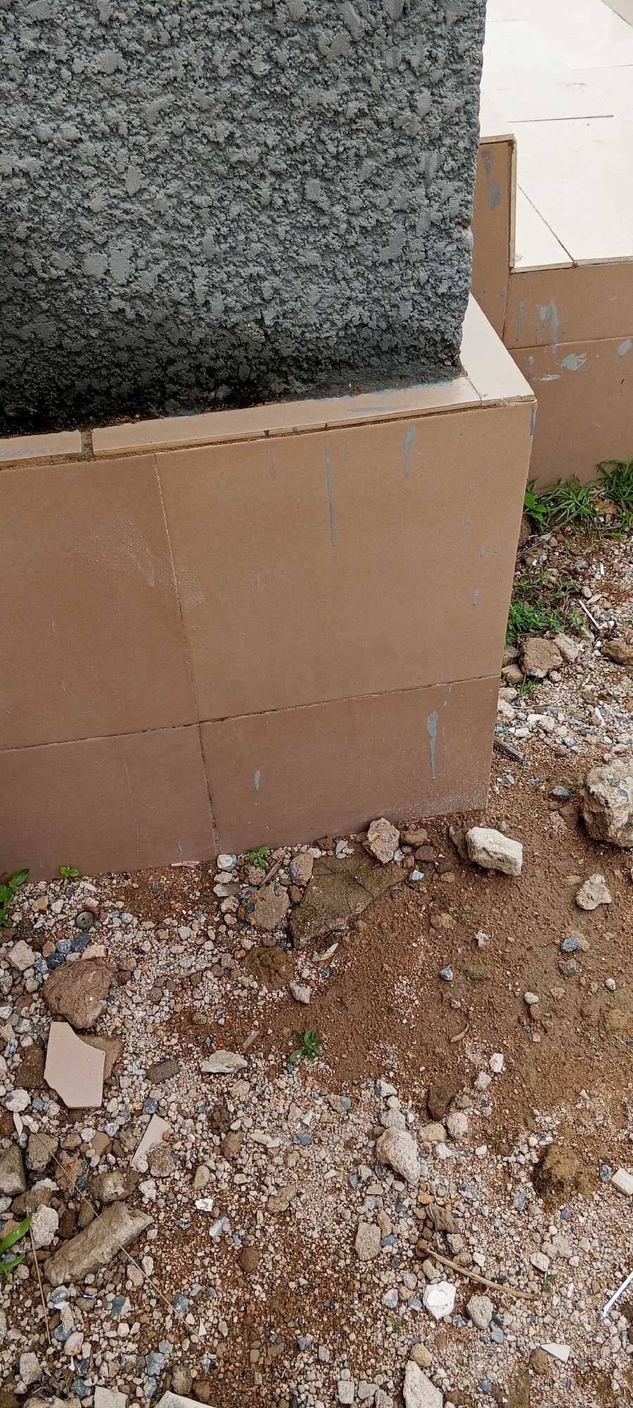 A close-up, eye-level shot shows the base of a rough, grey concrete structure adjacent to a wall tiled with brown, rectangular tiles. The ground in front of the tiled wall is covered in dirt, small stones, and debris, including broken pieces of concrete and tile. Sparse green weeds sprout from the ground. To the right of the tiled wall, a portion of a beige tiled surface is visible, suggesting a raised area or step. The scene appears to be outdoors in an unkempt urban or construction area.