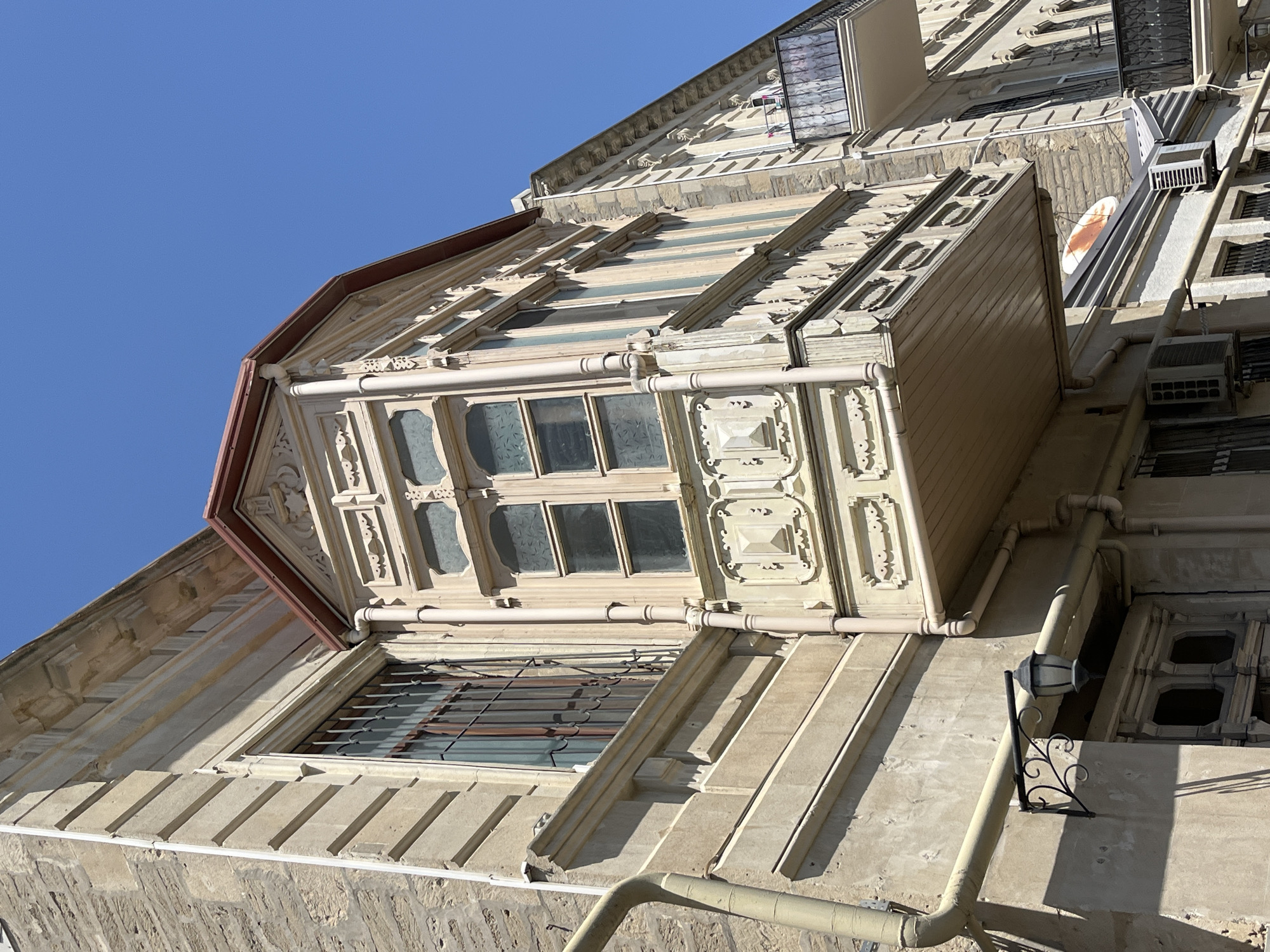 A close-up, low-angle outdoor shot captures the facade of an aged, light-colored building against a clear blue sky. The building exhibits intricate architectural details, including decorative molding, a bay window with multiple panes, and a wrought-iron balcony on an adjacent building. A yellow pipe runs vertically along the left side of the building, and a black wrought-iron lantern with a decorative scroll arm is mounted on the wall near a window. The lower part of the building shows air conditioning units and a small satellite dish. The overall scene suggests an urban environment with older architecture.
