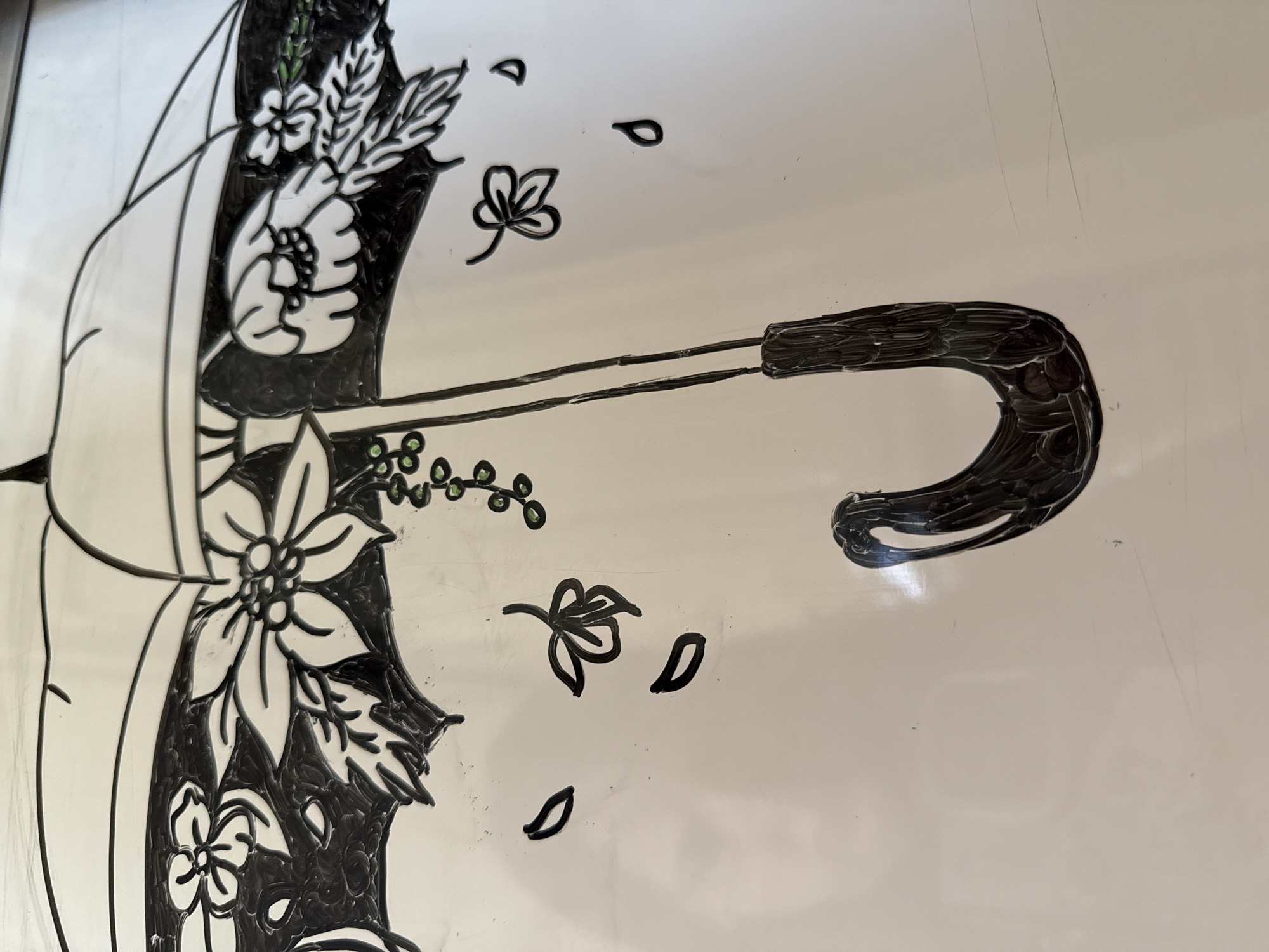 This media file displays a decorative illustration of an umbrella, partially visible on a plain, light-colored background. The umbrella is adorned with intricate floral and leaf patterns, rendered primarily in black with white outlines, and features small green accents for berries. The handle of the umbrella is prominently shown on the right side of the image, characterized by a dark, curved design with textured brushstrokes. Scattered around the main umbrella design are individual black-outlined leaves and small clusters of green berries. The overall aesthetic suggests a stencil-like or hand-painted design, possibly on a glass surface or a whiteboard, as indicated by faint linear scratches visible in the background. There are no people, explicit activities, specific time of day, weather conditions, or visible text in the image.