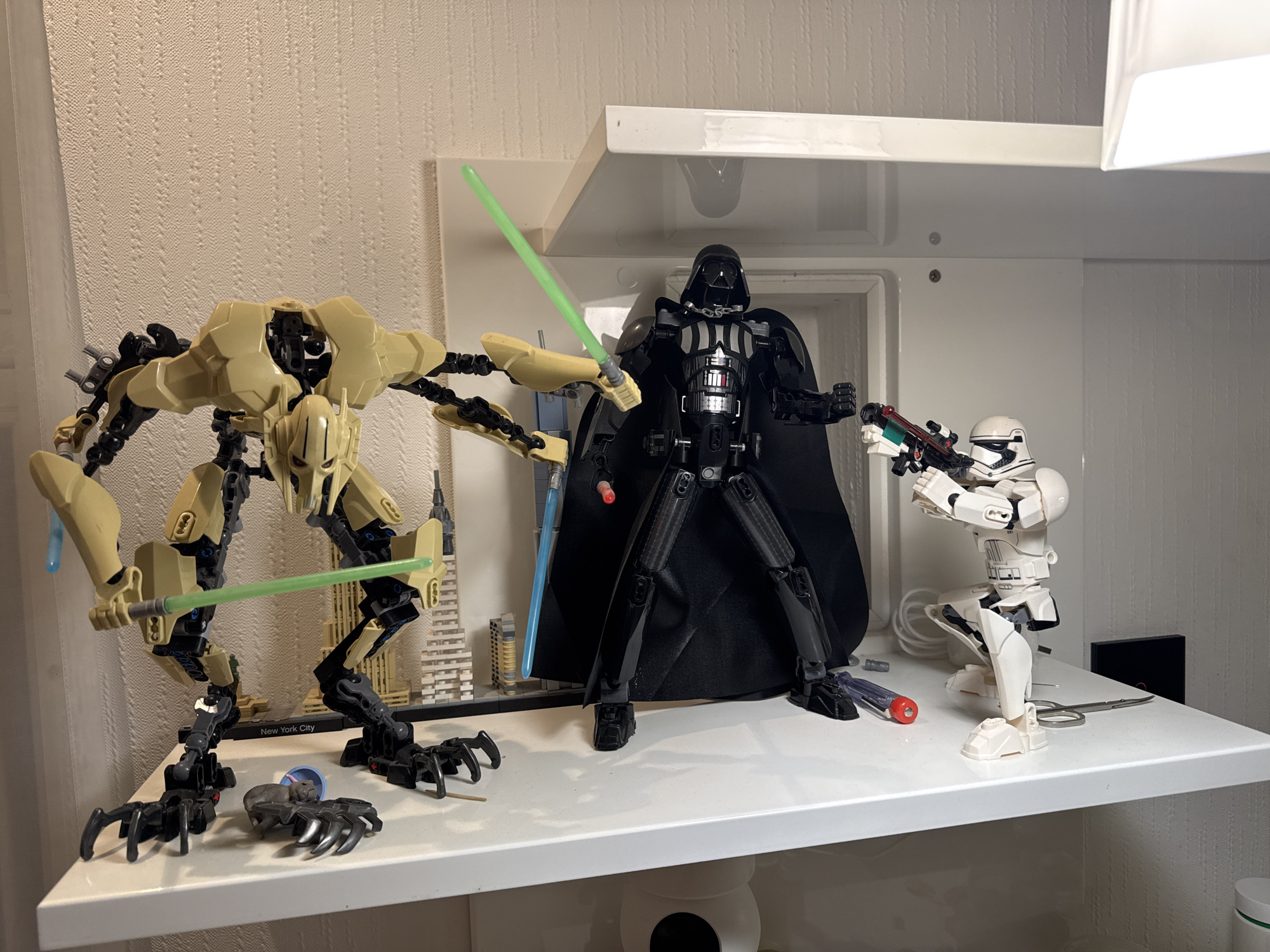 On a white shelf, a collection of Star Wars Lego figures are arranged. General Grievous, wielding two green lightsabers, stands to the left. Darth Vader, cloaked and also wielding a lightsaber, is positioned in the center. A Stormtrooper is to the right, holding a blaster rifle. In the background, a Lego model of the New York City skyline is visible. Additional tools and small items are scattered on the shelf around the figures. The setting appears to be a domestic environment, with textured wallpaper and a white shelving unit. There is no indication of the city of Volzhsky, Russia in this image.