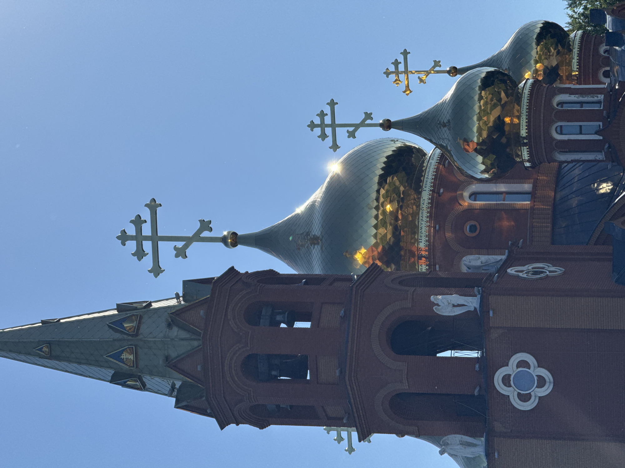 The image captures the spires and domes of the Trinity Cathedral in Chaikovsky, Russia, against a clear blue sky. The cathedral features a tall, pointed red brick tower with bells visible through arched openings. This tower is adorned with decorative white clover-shaped elements and statues of angels. Adjacent to the tower are multiple golden domes, some reflecting the sunlight, each topped with an Orthodox cross. The architecture suggests a traditional Russian Orthodox church.