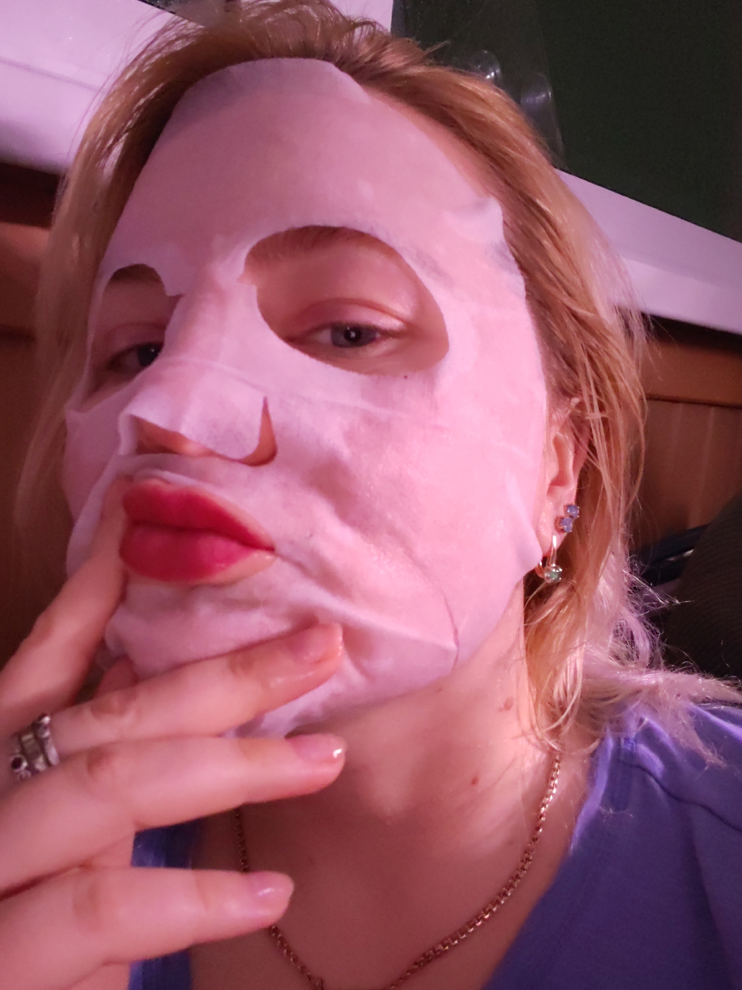 A fair-skinned person with blond hair is posing for a selfie indoors. A sheet mask, which is pink-hued, covers the majority of the person's face, with cutouts for the eyes, nostrils, and mouth. The lips are visible through the mouth cutout and are painted with a bright pink or red lipstick. The person is touching the mask with their left hand, which is adorned with a silver ring with dark-colored stones. They are wearing dangling earrings with blue and green stones. A gold chain necklace is visible around the person's neck, and they are wearing a purple shirt. There is no information to suggest this image was taken in Izhevsk, Russia.