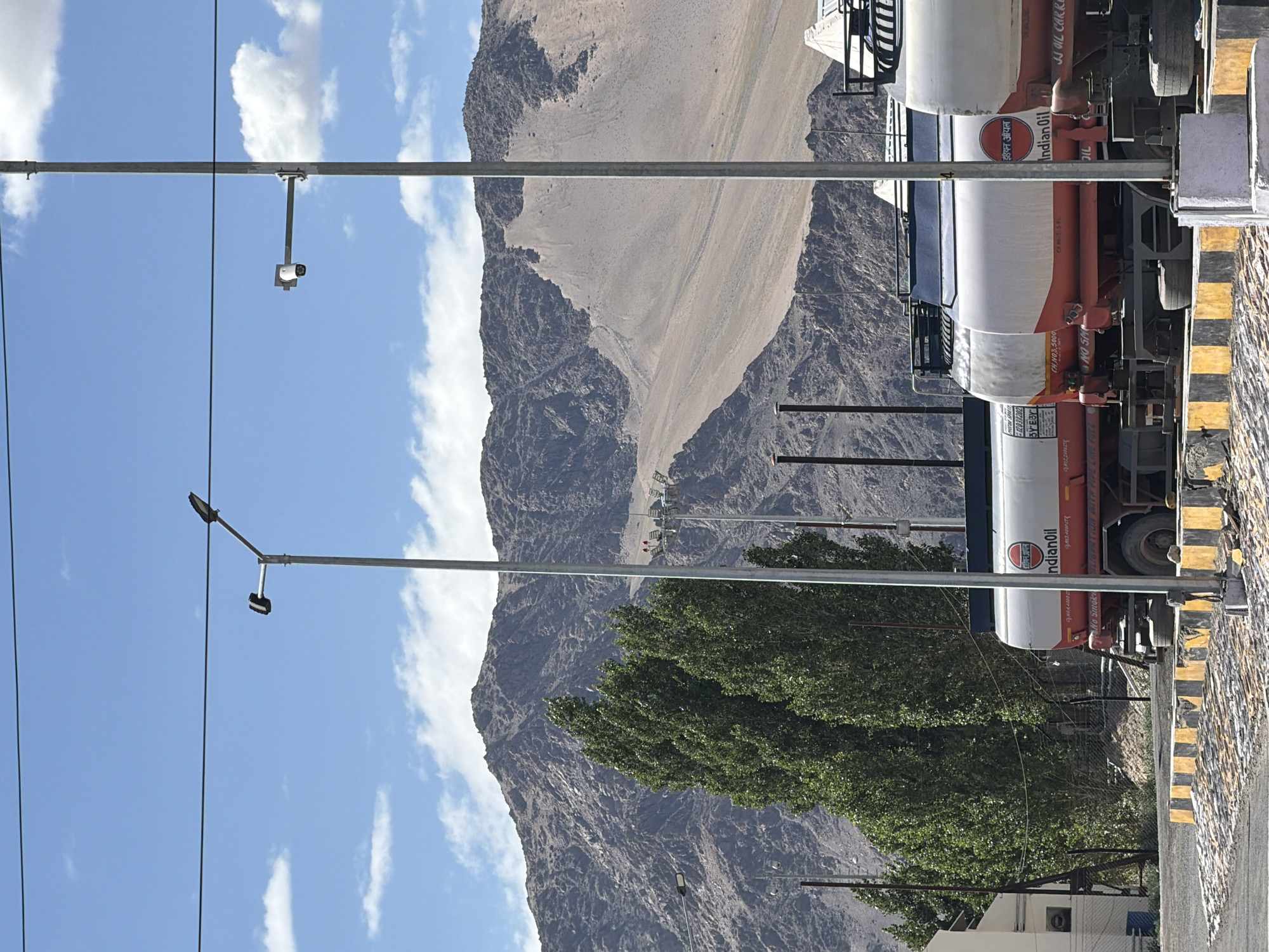 The image depicts a clear, sunny day in the city of Leh, India, with a mountainous landscape in the background. In the foreground, several fuel tankers with "IndianOil" branding are parked. The tankers are white and orange, and some have visible text including "No Smoke" and "Oil". A yellow and black striped barrier is present in front of the tankers. Tall, green trees are situated to the left of the tankers, and a building with a white exterior is partially visible behind them. Two tall, metallic lampposts with lights and a security camera are prominent in the scene. Power lines are stretched across the top of the frame. The mountainous terrain is characterized by a mix of rocky slopes and lighter-colored sediment. The sky is blue with scattered white clouds. There are no people visible in the image.