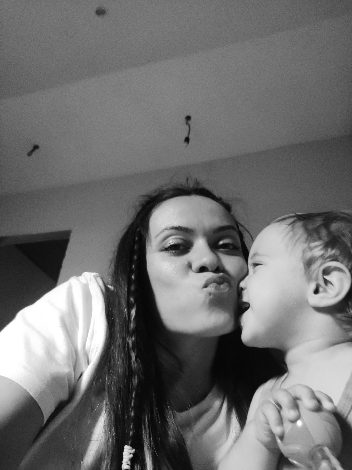 This black and white, low-angle, medium-shot photograph captures a mother and child sharing a tender moment indoors. The woman, with long dark hair, is on the left, looking towards the camera with her lips puckered in a kiss gesture. She is wearing a light-colored, possibly white, short-sleeved top. Her eyes are visible, and she has visible eyelashes and possibly eyeliner.
To her right, slightly in front of her, is a young child, possibly a toddler, facing away from the camera and towards the woman's face, with an open mouth and appearing to be about to kiss or make a sound. The child has light-colored hair and is wearing what appears to be a light-colored top or strap. The child's right hand is visible, holding a bottle with a slightly darker cap or spout.
The background is a simple, light-colored wall or ceiling, possibly indicating an indoor setting. There are a few indistinct dark spots on the wall/ceiling in the upper left and upper middle of the frame, which might be light fixtures or imperfections. The lighting appears to be somewhat dim, contributing to the intimate feel of the image. The overall mood is one of affection and closeness between the two subjects. No visible text is present in the image.