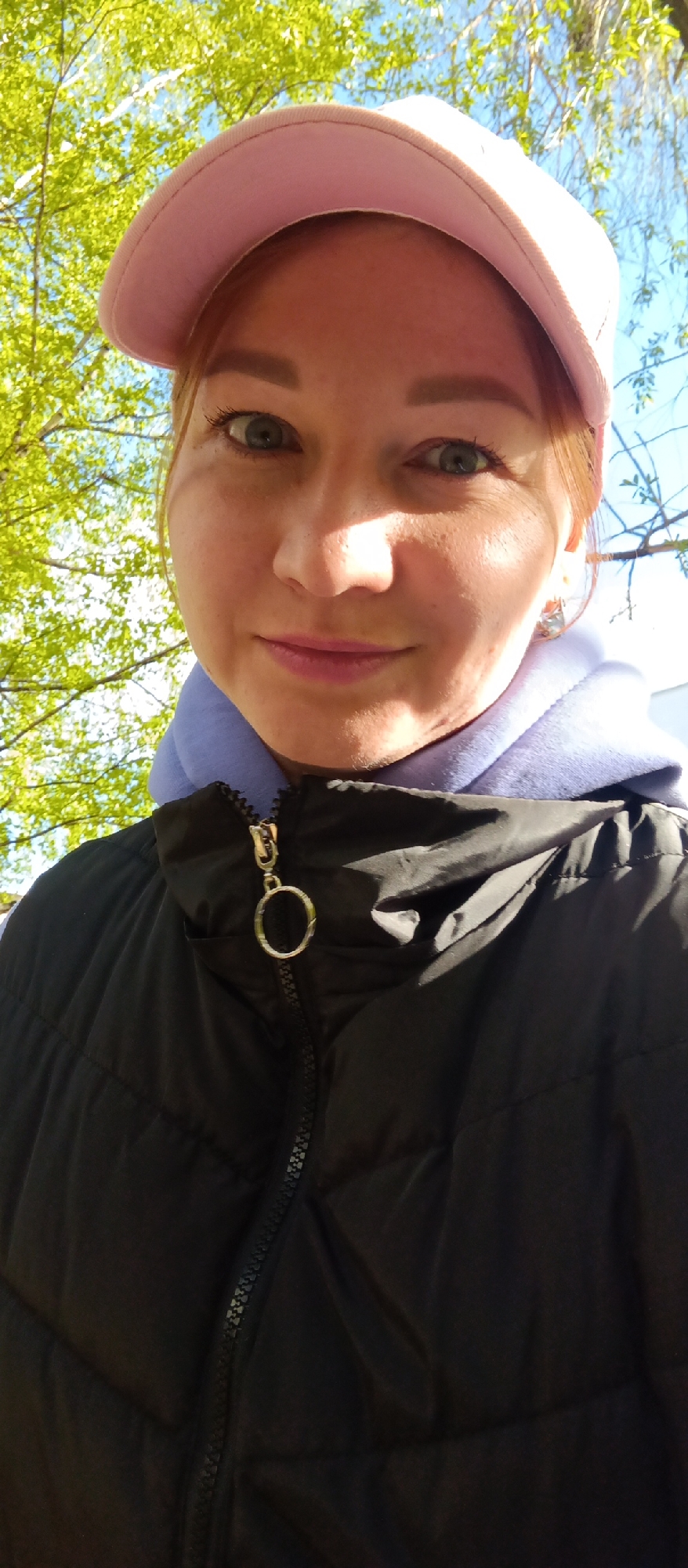 A woman with reddish hair wearing a pink baseball cap and a black puffy jacket over a light purple hooded sweatshirt stands outdoors. The zipper of the jacket is partially open, revealing the sweatshirt. The background includes green foliage from trees and a glimpse of a blue sky. The location appears to be outdoors in an area with trees.