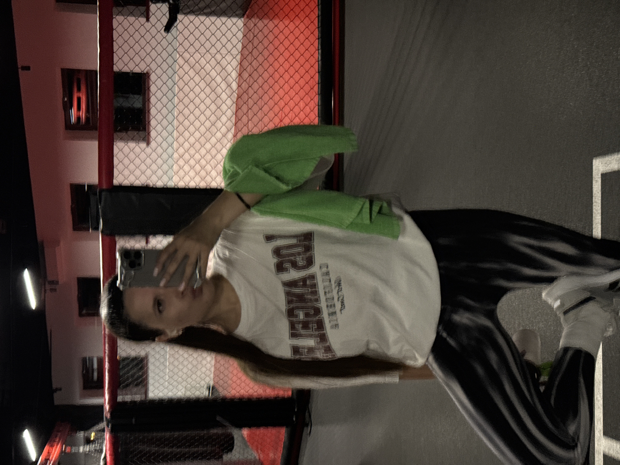 A woman is taking a selfie in what appears to be a gym or workout facility. She is wearing a white t-shirt with "LOS ANGELES" printed on it, tie-dye leggings, and white sneakers. A green towel is draped over her shoulder. The background includes a fence-like structure with red framing and a red wall with windows. The location cannot be determined to be Vniissok, Russia, from the image.