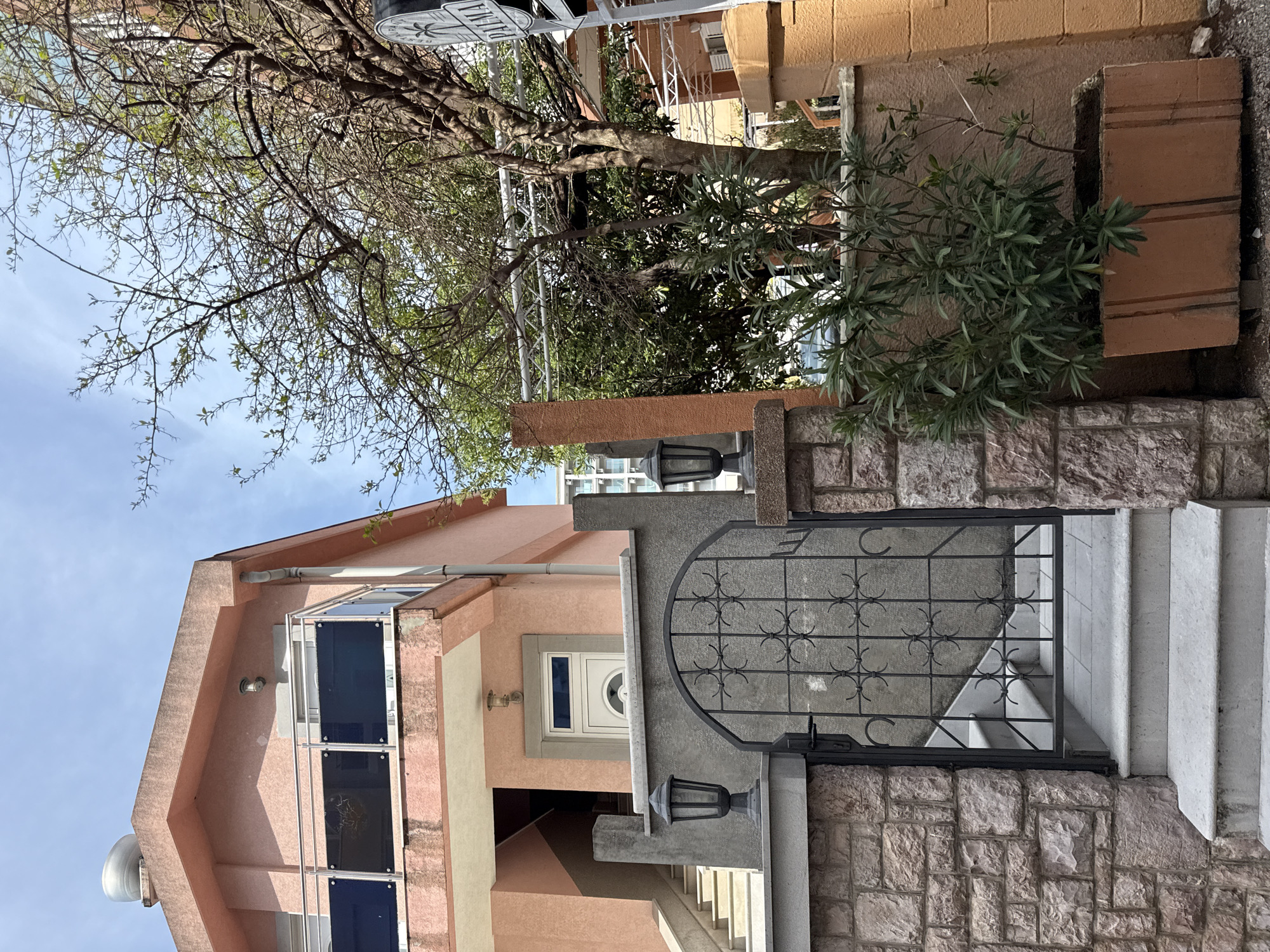 The image shows a street scene in an urban area, with a building prominently featuring a metal gate leading to a set of stairs. The building has a pink facade with multiple windows. A tree overhangs the structure, partially obscuring the sky. A plant sits beside the gate on a stone wall. The sky visible is partly clear with a blue hue. The location is Unknown, Unknown.