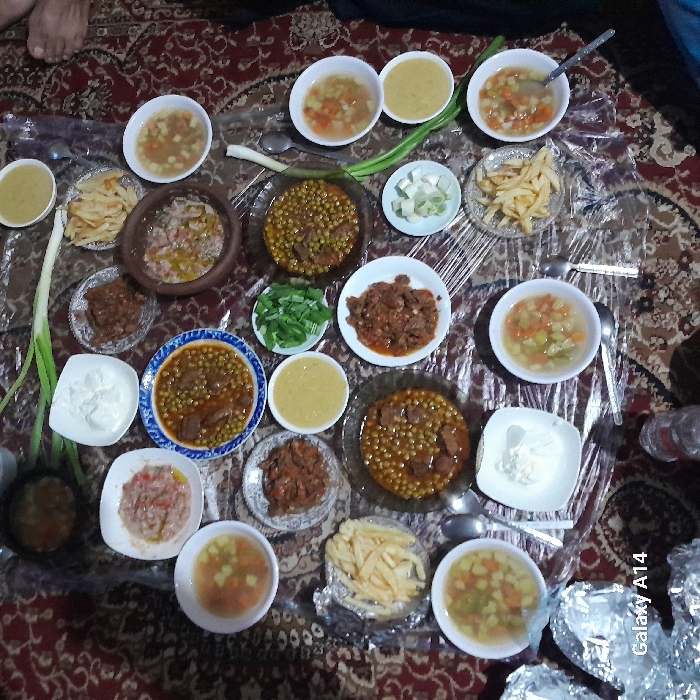 This image depicts a communal meal arranged on a floor in a domestic setting within the city of Khan Yunis, Palestinian Territory. The meal is spread across a clear plastic sheet overlaying a patterned, reddish-brown carpet. The scene, captured from an overhead perspective, displays a variety of dishes. These include at least six white bowls containing vegetable soup with diced carrots and potatoes, three large plates or bowls of a meat and pea stew, and approximately five smaller plates of a reddish-brown meat stew. Additional items include three bowls of a yellow puréed soup (likely lentil), two bowls of a white creamy substance (likely yogurt or labneh), two plates of french fries, one plate of finely chopped mixed fresh vegetables, and small bowls containing chopped green and white onions. Several whole green onion stalks are scattered directly on the plastic sheet. A dark, ceramic pot containing a cooked dish is also visible. Spoons are present in most soup bowls and several stew dishes. Portions of two bare human feet are visible on the carpet at the right side of the frame, indicating the presence of at least two individuals seated on the floor for the meal. A transparent plastic bottle is partially visible at the bottom center, and several foil-wrapped bundles are in the bottom left corner. The image includes a "Galaxy A14" watermark.