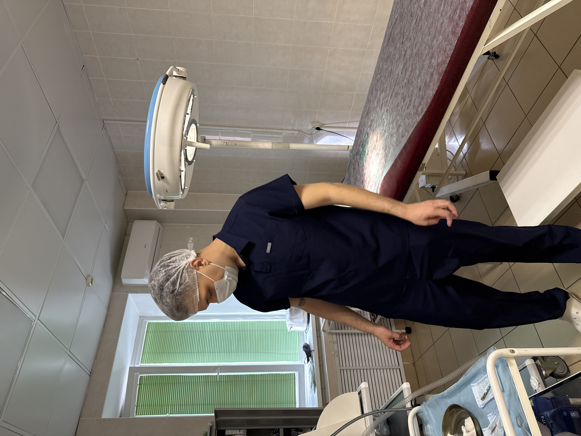 The image shows a man in a dark blue surgical scrub suit, a white surgical cap, and a white face mask, standing in what appears to be a medical examination room or operating theater. He is looking down, presumably at something below the frame.
The room is equipped with a surgical light fixture mounted on the ceiling, a window with green blinds, and a medical examination table. Various medical equipment and supplies are also visible, including a cart with a metal bowl containing surgical tools.
While the location in Perm, Russia is unconfirmed, the setting suggests a medical facility based on the presence of surgical equipment and clothing.