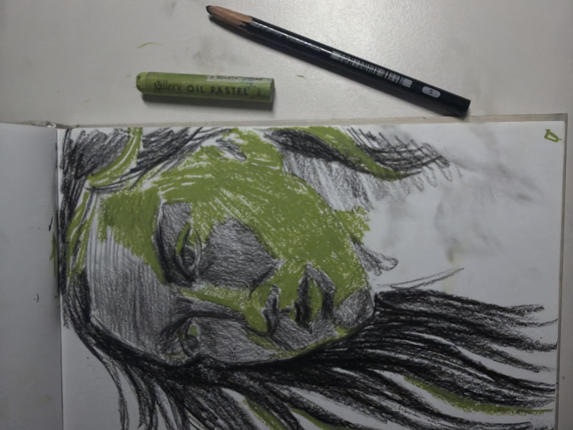 An unfinished portrait sketch of a person's face is shown, rendered in charcoal and a vibrant green oil pastel. The sketch is on white paper, with charcoal used for shading and defining features, and the green pastel applied to highlight sections of the face and hair. To the right of the sketch, a green oil pastel stick and a black pencil are placed on a white surface. The scene appears to be an artist's workspace. The image does not contain any recognizable landmarks or elements that would allow for the identification of its location within Dongducheon-si, South Korea.