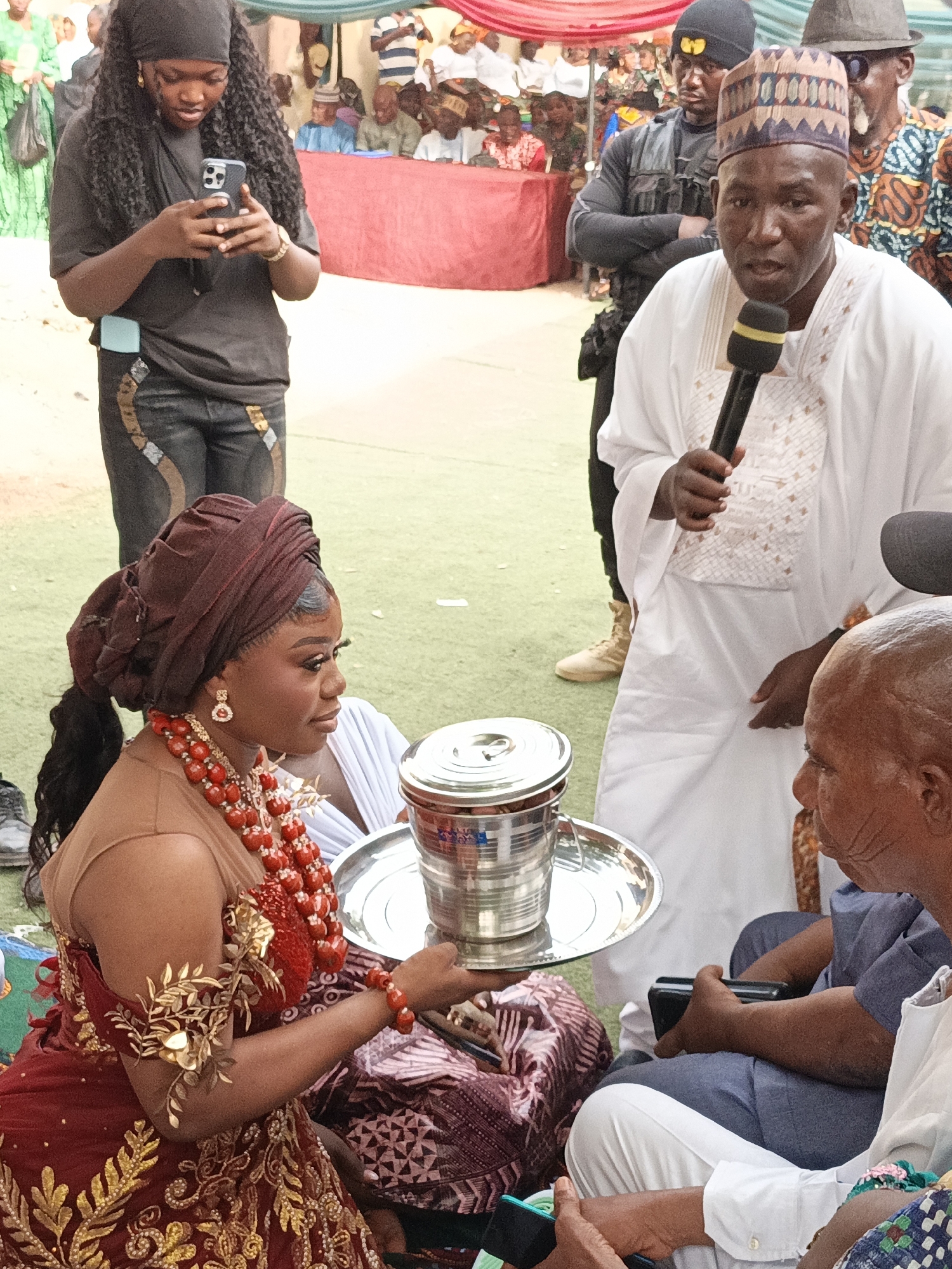 This image captures a vibrant traditional ceremony, likely a wedding or engagement, unfolding outdoors in Sabon Gari West, Nigeria.
In the foreground, a young woman, presumably the bride, sits elegantly, dressed in elaborate traditional attire. She wears a rich maroon headwrap, a top with sheer detailing, and a floor-length gown adorned with intricate gold patterns on a dark red fabric. Around her neck is a prominent, multi-layered red beaded necklace, complemented by matching earrings and a golden, leaf-inspired armlet. She is holding a large, reflective silver tray on which a lidded, cylindrical metal container rests, suggesting an offering or an item central to the ritual.
To her right, an older, bald man, wearing a white garment and blue trousers, is seated on the ground, observing the scene. In the midground, a man in a white traditional robe and a patterned cap addresses the attendees, speaking into a black microphone. Behind him, a man in a black tactical vest and beanie stands guard. To the left, a young woman with long, curly hair and a black headscarf stands, engrossed in her smartphone. She wears a black t-shirt and dark jeans with unique gold-colored accents.
The setting features a bright green artificial turf ground and a red canopy providing shade. Numerous guests are visible in the background, seated and standing under the canopy, many dressed in traditional attire, indicating a significant social gathering. The event is taking place during daylight hours, under clear and bright conditions. There is no visible text in the image.