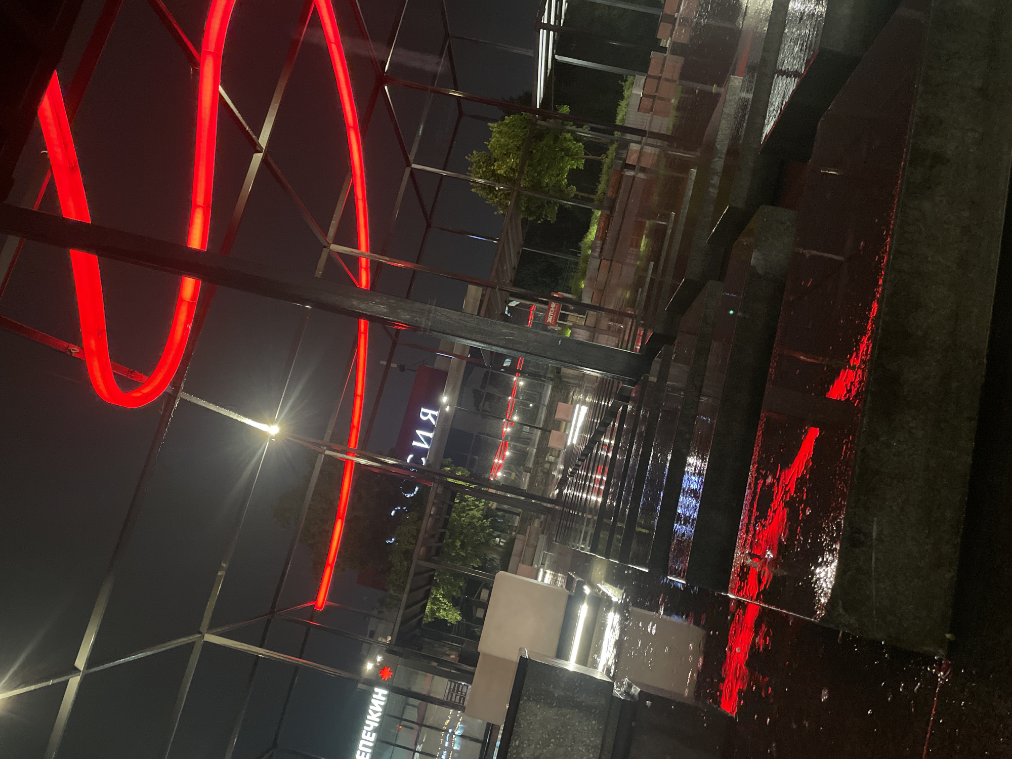 The image depicts a nighttime scene in Izhevsk, Russia, characterized by wet surfaces reflecting artificial light. A prominent architectural feature is a red neon structure composed of curved and straight lines, forming a partial enclosure or frame. The reflective surfaces indicate recent rainfall or a wet environment, amplifying the illumination from streetlights and neon signs. In the background, buildings with illuminated signs in Cyrillic script are visible, suggesting a commercial area. The composition includes elements of metal framework, possibly part of the structure or nearby construction. The perspective is from a low angle, emphasizing the height and scale of the scene. The overall atmosphere is urban and modern.