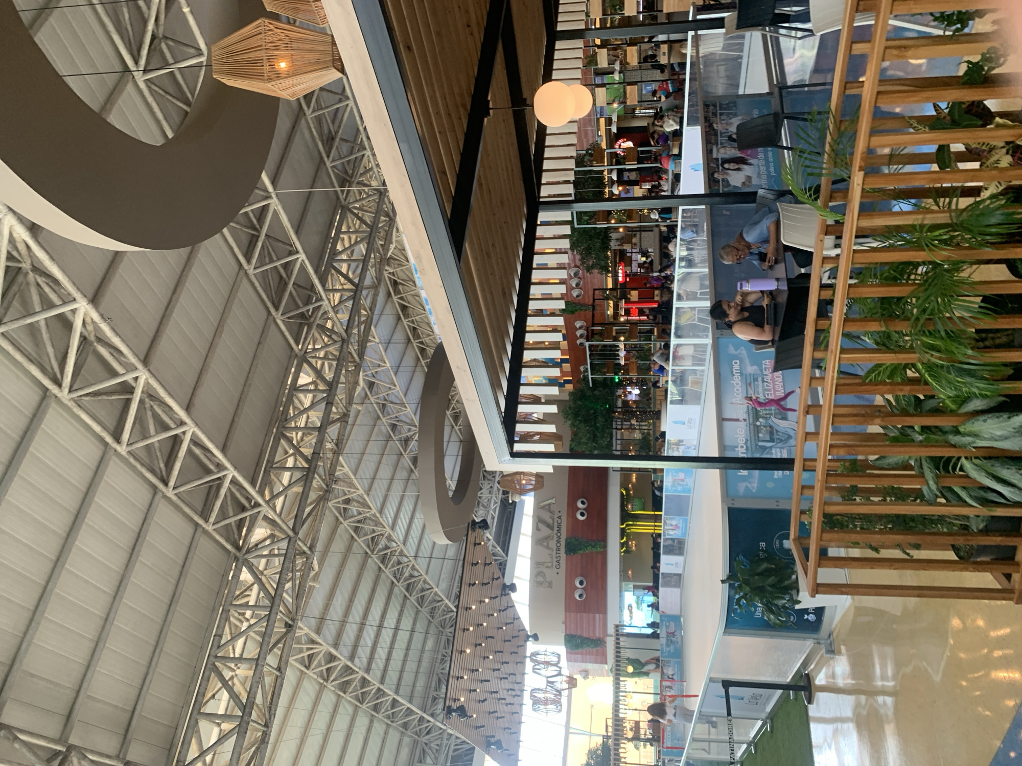 This image depicts an indoor public space, likely a mall or entertainment complex, in Porlamar, Venezuela. A prominent feature is an ice skating rink with blue and white barriers, on which a lone individual is visible skating. Surrounding the rink are various commercial establishments, including an area labeled "PLAZA GASTRONOMICA" with distinctive cartoonish eyes above its entrance. Several people are seated at tables in an outdoor seating area in the foreground, with two women engaged in conversation and looking at a tablet. The architecture features a high, exposed metal roof structure and modern lighting fixtures, including large pendant lamps. Green plants are visible in planters and along a wooden railing in the foreground, adding a touch of nature to the urban environment.
