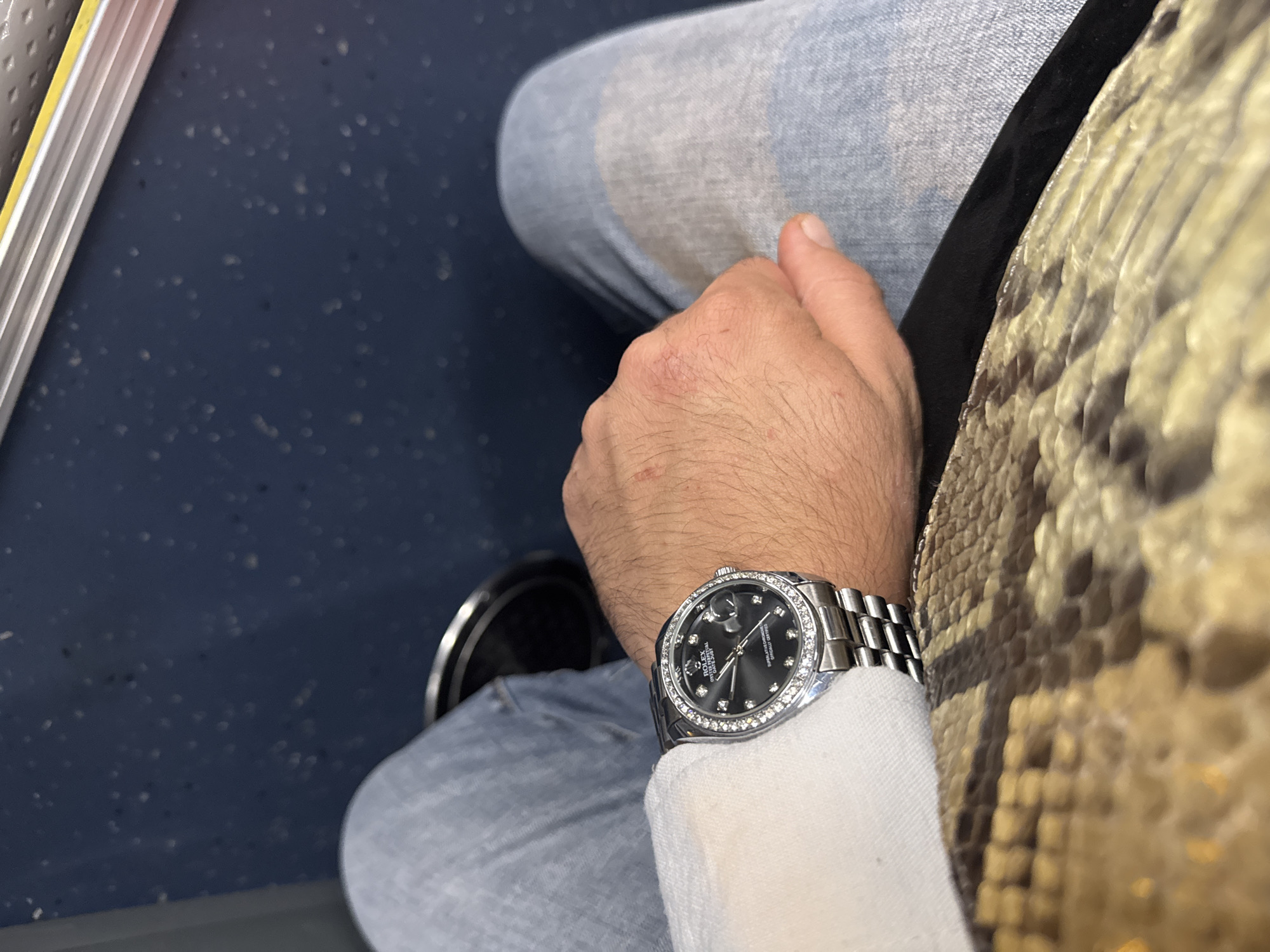 A high-angle shot depicts a person seated, likely on public transport due to the blue, textured surface of the seat and the presence of a metallic bar along the left edge. The person is wearing light blue jeans and a silver-tone Rolex watch with a diamond bezel. Their right hand rests near their lap. A portion of a snakeskin-patterned item is visible on the right side of the frame. The location is indeterminate; Yerevan, Armenia cannot be ascertained from the image.