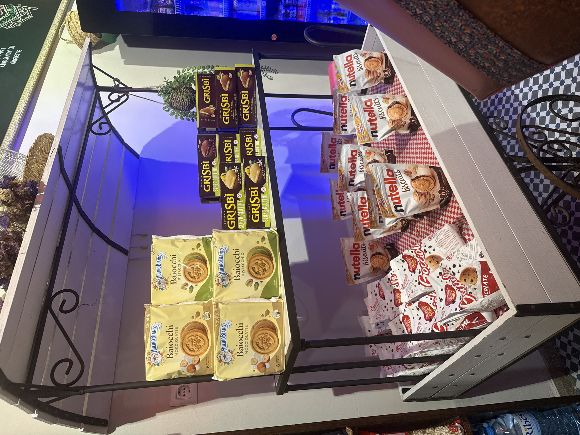 This image captures a retail display in Tossa de Mar, Spain, featuring a variety of packaged biscuits and snacks. The scene is set in an indoor environment, likely a shop or cafe, characterized by a rustic wooden cart with multiple shelves.
On the upper shelves, several yellow packages of "Mulino Bianco Baiocchi" biscuits are prominently displayed, in both "Nocciolatte" (hazelnut) and "Pistacchio" (pistachio) flavors. Next to these, stacked boxes of "Grisbì" biscuits, in "Cioccolato," "Limone," and "Pistacchio" flavors, are arranged in a pyramid formation. Some of the Grisbì boxes are labeled "Senza Glutine" (gluten-free).
The lower section of the cart contains a white wooden bin filled with more packaged snacks. Most visible are numerous white pouches of "Nutella Biscuits." Interspersed with these are bags of "Gocciole" biscuits, a popular Italian brand. A person's arm and shoulder, wearing a patterned sleeve, are visible on the right side of the frame, suggesting their presence near the display.
The background shows a wall with some decorative elements, including a woven basket and some dried floral arrangements. A vending machine is partially visible on the far right. The lighting in the scene is a mix of ambient light and possibly colored LED lighting, creating a purplish hue on the wall. The floor has a distinctive black and white checkered tile pattern. The overall impression is of a well-stocked confectionery display in a quaint Spanish establishment.
