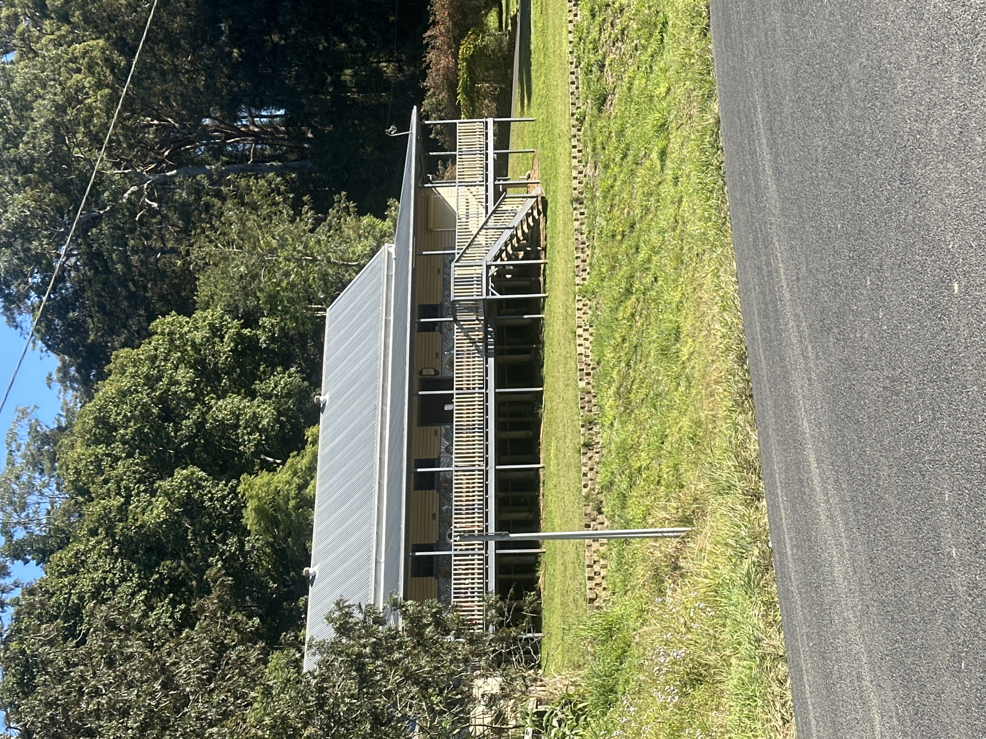 The image shows a light-colored wooden house with a corrugated metal roof, situated on stilts in a grassy, rural area. The house has a wraparound porch with white railings and a set of stairs leading up to it. A dense forest of tall trees forms the backdrop of the scene. The foreground features a paved road. The scene is bathed in sunlight, casting shadows on the ground and highlighting the natural environment. There are no people or animals visible in the image. The location is in Thora, Australia, known for its lush, natural surroundings and rural setting.