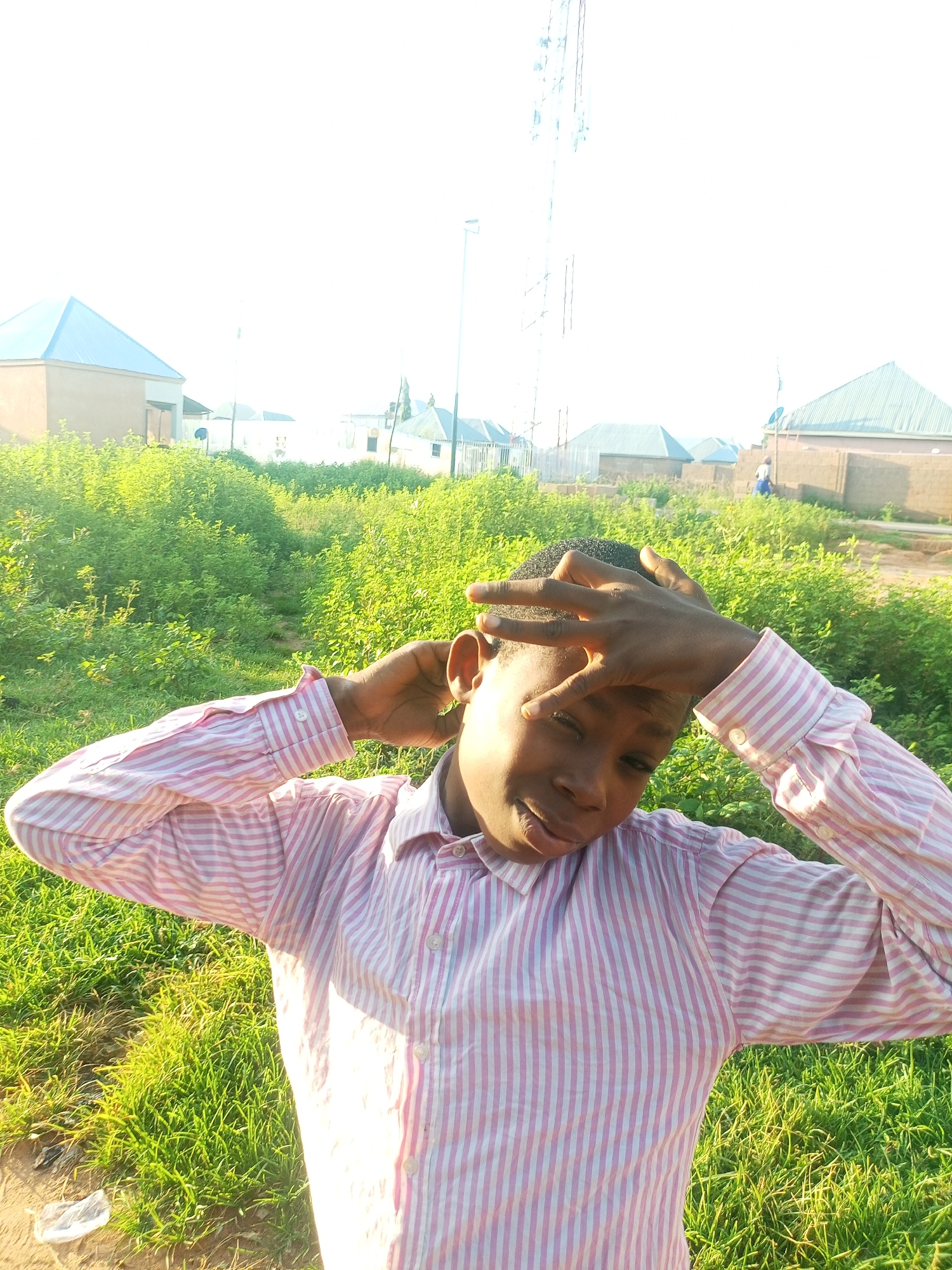 A young Black male is pictured in a close-up, medium shot. He is outdoors in what appears to be a semi-urban or rural area of Tempure, Nigeria. The subject is wearing a pink and white striped collared shirt. He has his left hand on the back of his head and his right hand touching his forehead. His head is slightly tilted and he is looking towards the camera with a relaxed expression. The background consists of lush green vegetation and several buildings with light-colored walls and pitched roofs, typical of Nigerian architecture. A tall communication tower is visible in the distance. The lighting suggests it is daytime, possibly late afternoon, with the sun casting shadows and illuminating the scene. There are no other people in the immediate vicinity of the subject.