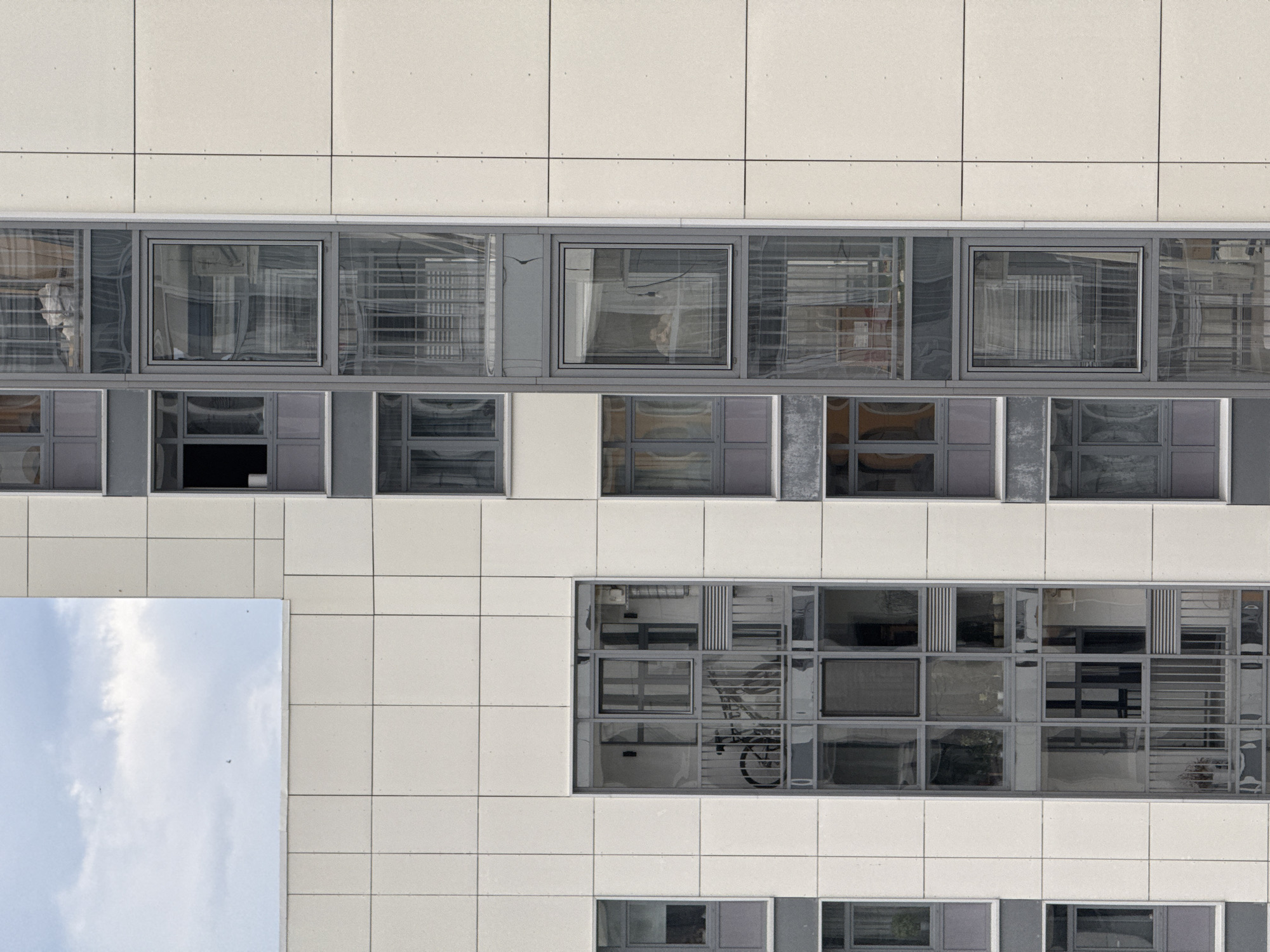 The image shows a section of a building exterior, potentially located in Perm, Russia, judging by the building style common in that area. The facade is composed of light-colored, square or rectangular panels arranged in a grid pattern. There are multiple rows of windows with gray frames. The windows reflect the surrounding environment, showing partial images of other buildings, and potentially a sky with clouds. One window shows a reflection of clouds. The reflections suggest a daytime setting with clear or partly cloudy weather. Some windows have items inside the rooms reflected in them, such as interior structures, objects and a bicycle. The overall impression is a modern or recently renovated building facade.
