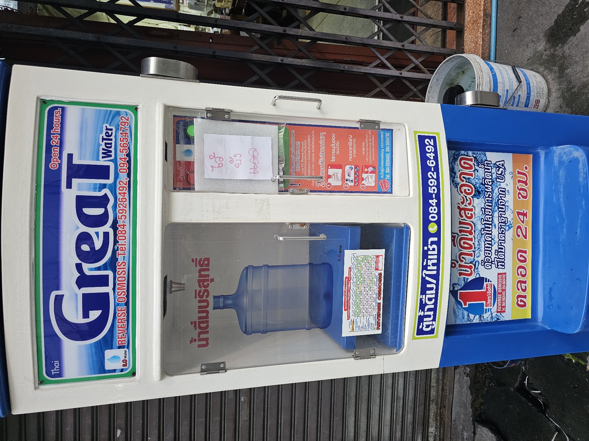 A self-service water dispenser machine is located on a street in Bangkok, Thailand. The machine has a blue and white exterior with the brand "Great Water" prominently displayed. It appears to be operating 24 hours a day. A large blue water bottle is visible inside the machine. The machine is positioned in front of a closed metal shutter door. A white bucket is placed to the right of the machine. The ground is concrete. The overall environment suggests an urban setting.