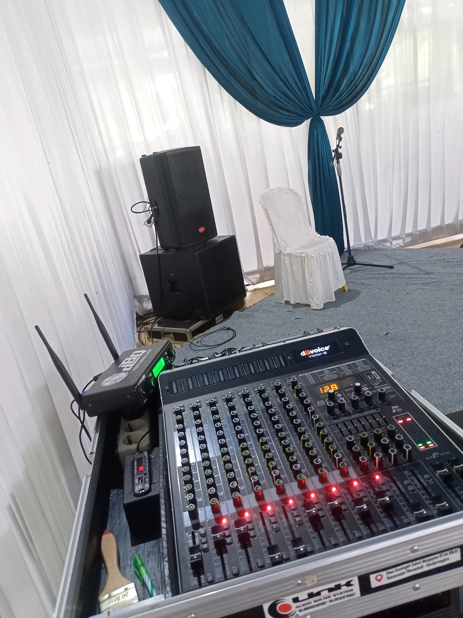 The image depicts a setup for a sound system, likely for an event, on a grey carpeted floor. In the foreground, a black audio mixer with numerous faders and knobs is prominently displayed. Red indicator lights are illuminated on several of the mixer's channels. To the left of the mixer, a black wireless receiver with two antennas sits on a raised surface. Behind the mixer, two black audio speakers are stacked, with the larger one on the bottom. Further back, a white chair draped in a ruffled fabric cover is positioned. A microphone on a stand is visible behind the chair, and a dark blue drape is arranged above the white curtains that form the backdrop. The bottom right corner of the image shows a label on the mixer case that reads "Desa Gembul Dukuh Margosari RT. 20 RW. 04 Kecamatan Wonodadi Banjarengara," indicating the location as Banjarengara, Wonodadi District, Margosari Hamlet, Gembul Village in Indonesia.